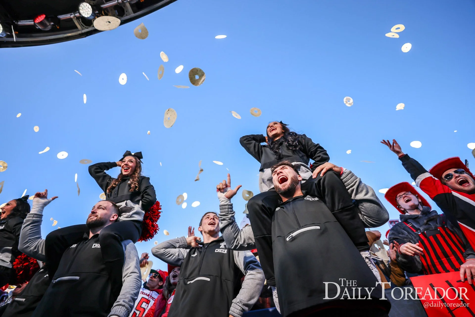 People celebrating at a sports event, with some on the shoulders of others, throwing confetti into the air, under a clear blue sky.
