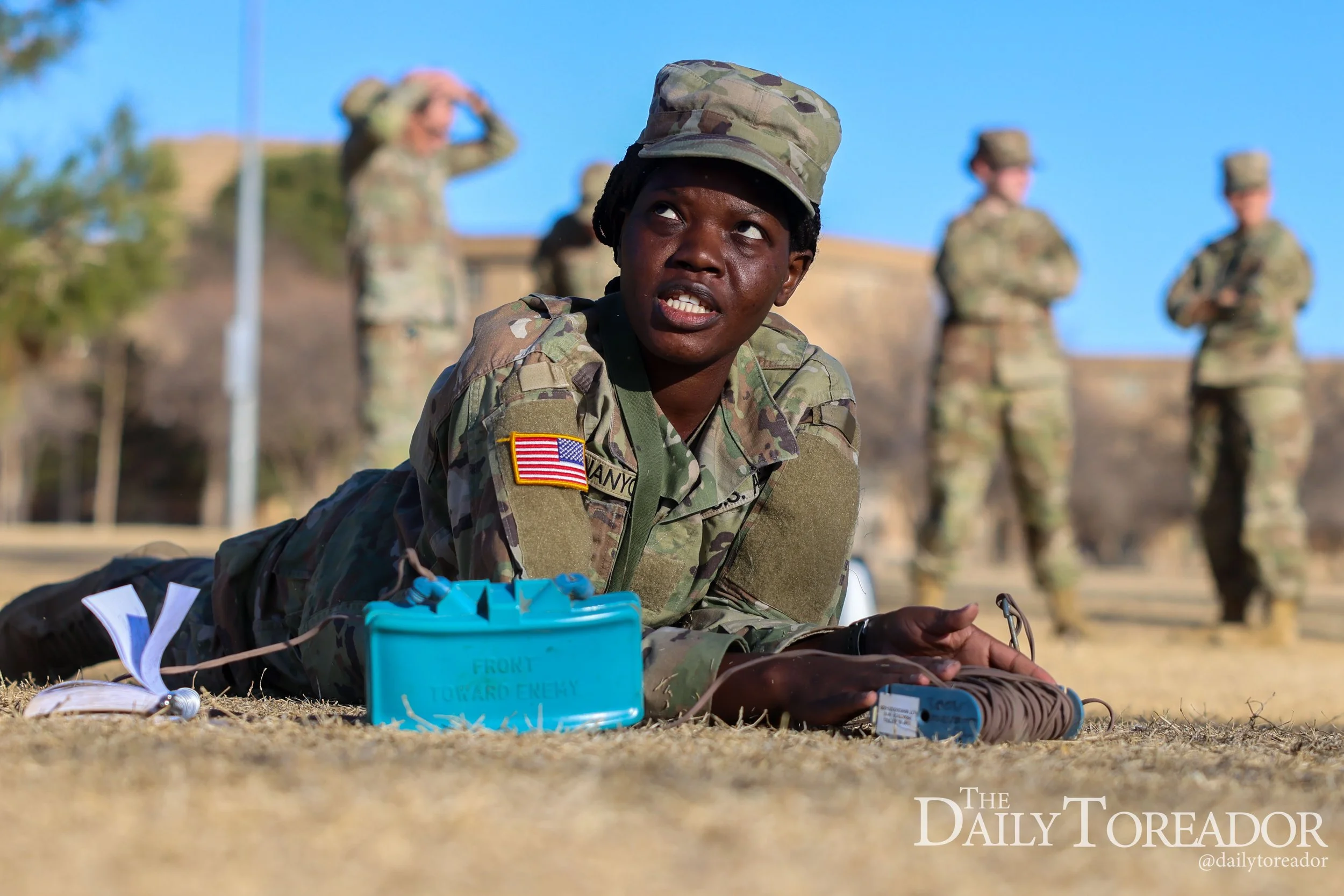 A soldier in camouflage uniform lying on the ground, surrounded by military equipment and supplies, with other soldiers standing in the background during daytime.