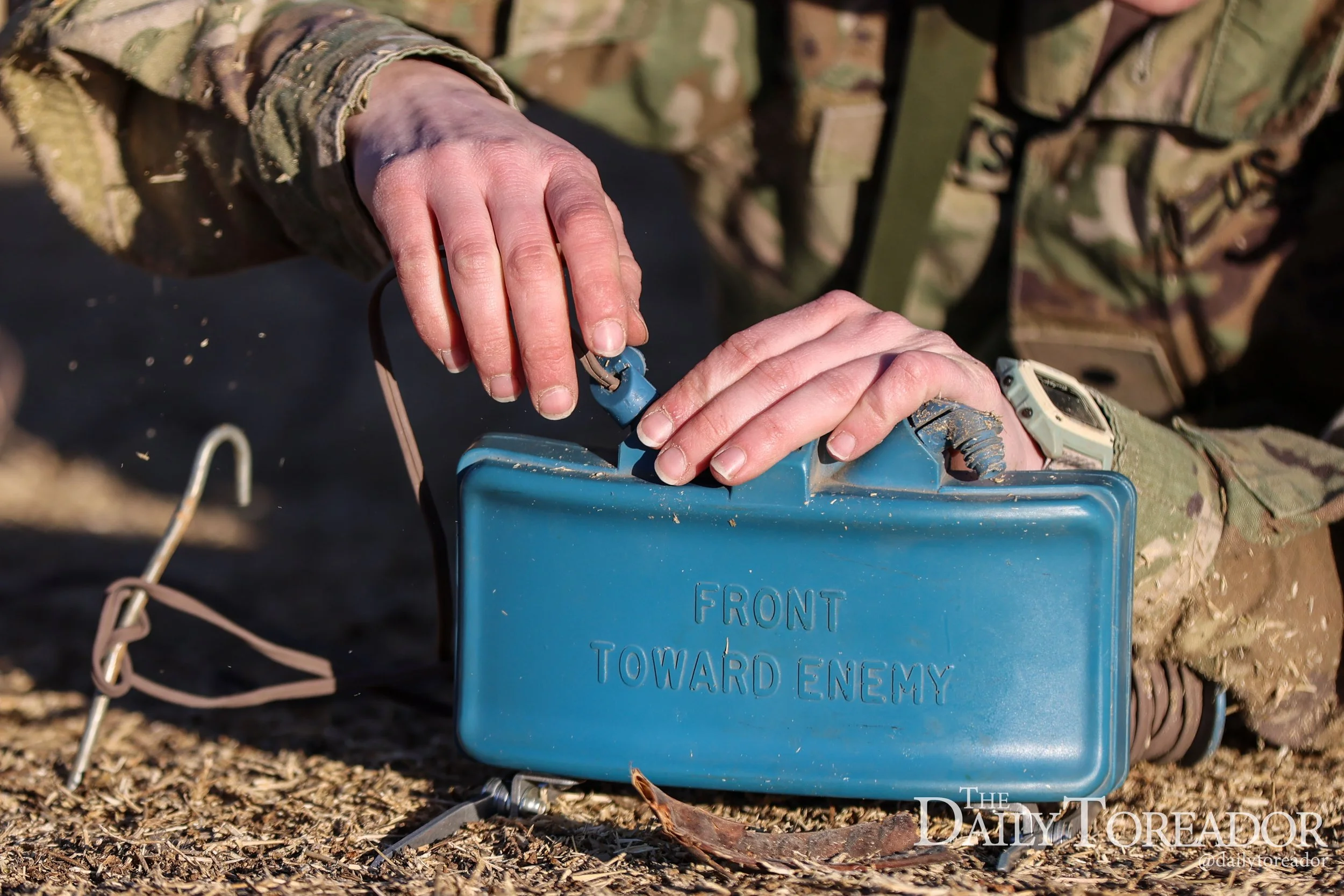 Close-up of a person in military camouflage placing a small blue box labeled "FRONT TOWARD ENEMY" on rough ground outdoors.