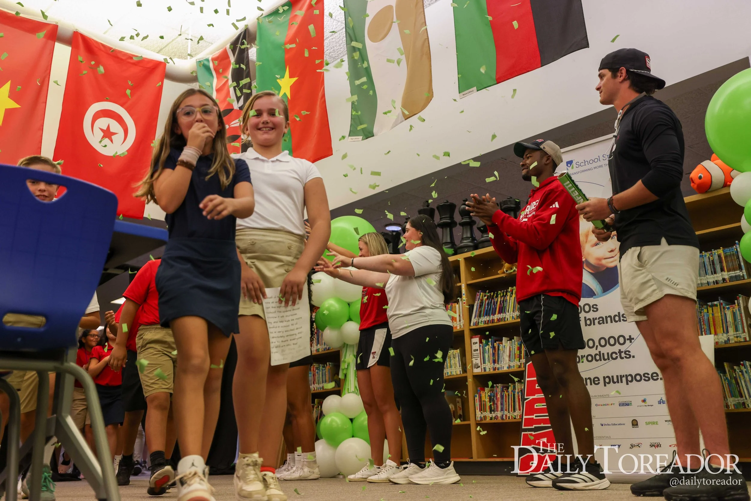Children and young adults celebrating in a library decorated with flags and balloons, with green confetti falling.