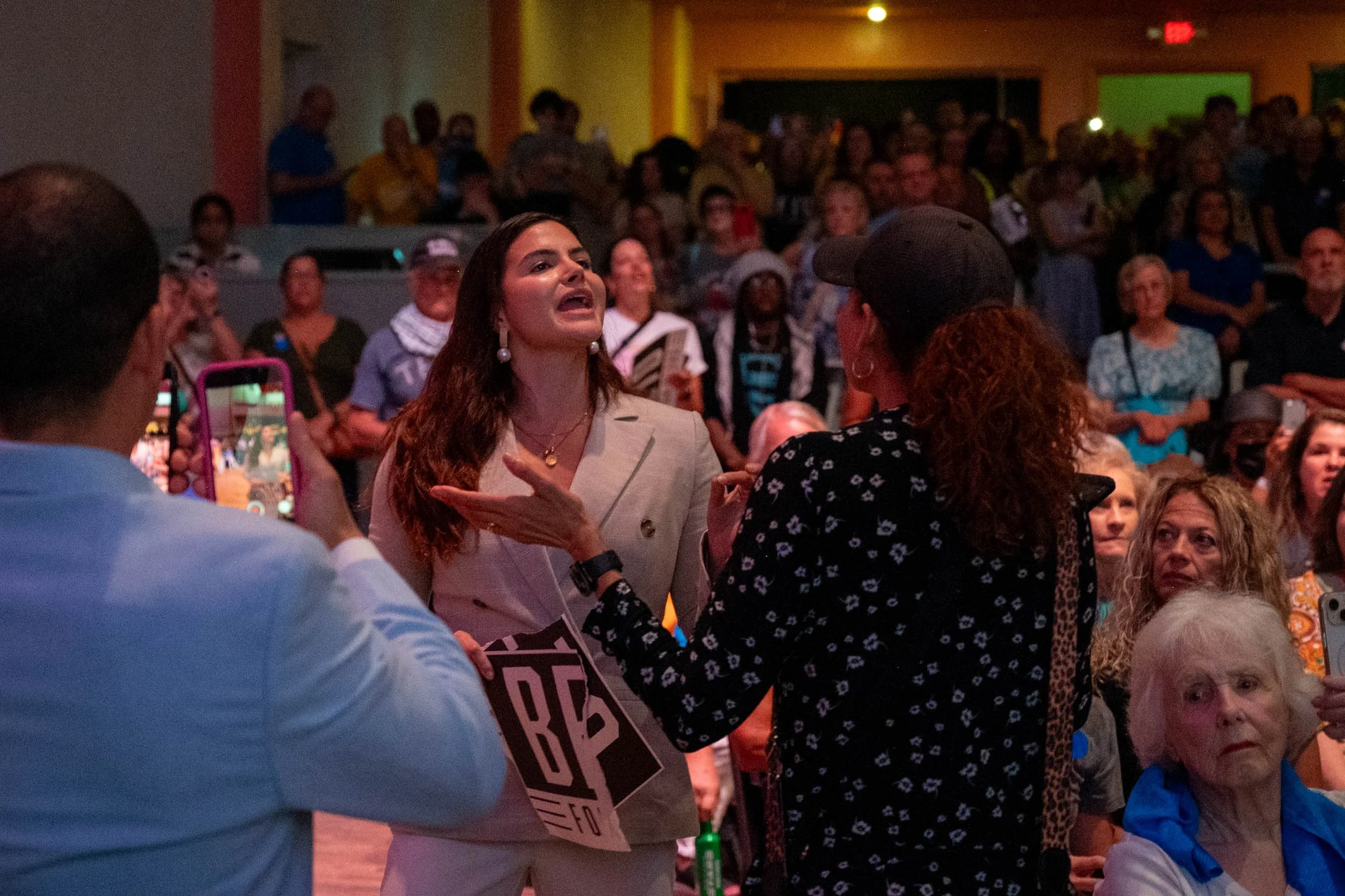 Two women are engaging in a conversation in front of a large crowd at an indoor event or rally, with several people taking photos or recording videos.