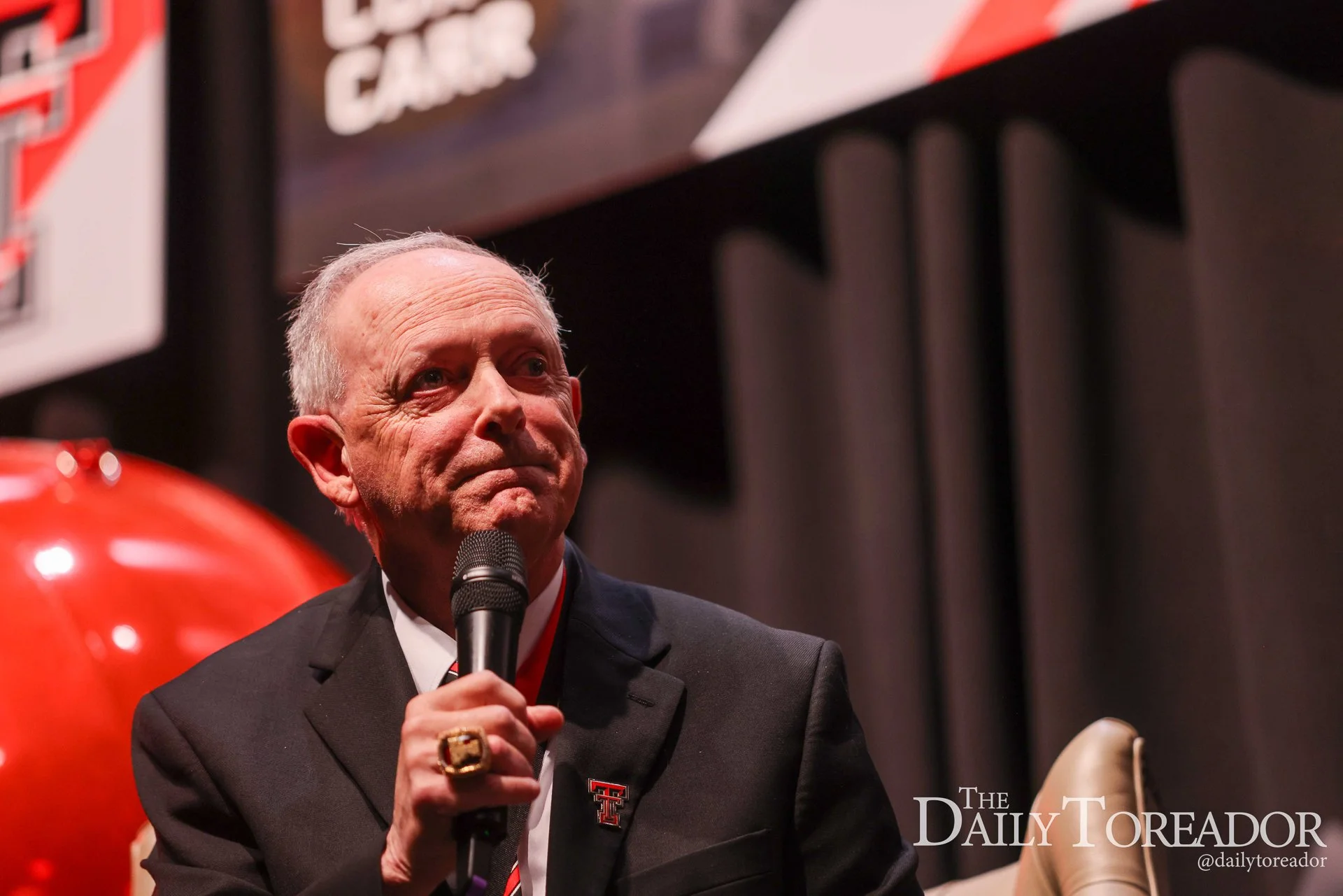 An older man with grey hair, wearing a black suit and a white shirt, is holding a microphone and speaking at an event. There is a red object in the foreground and a dark background with some partially visible signage and logos, including 'The Daily T