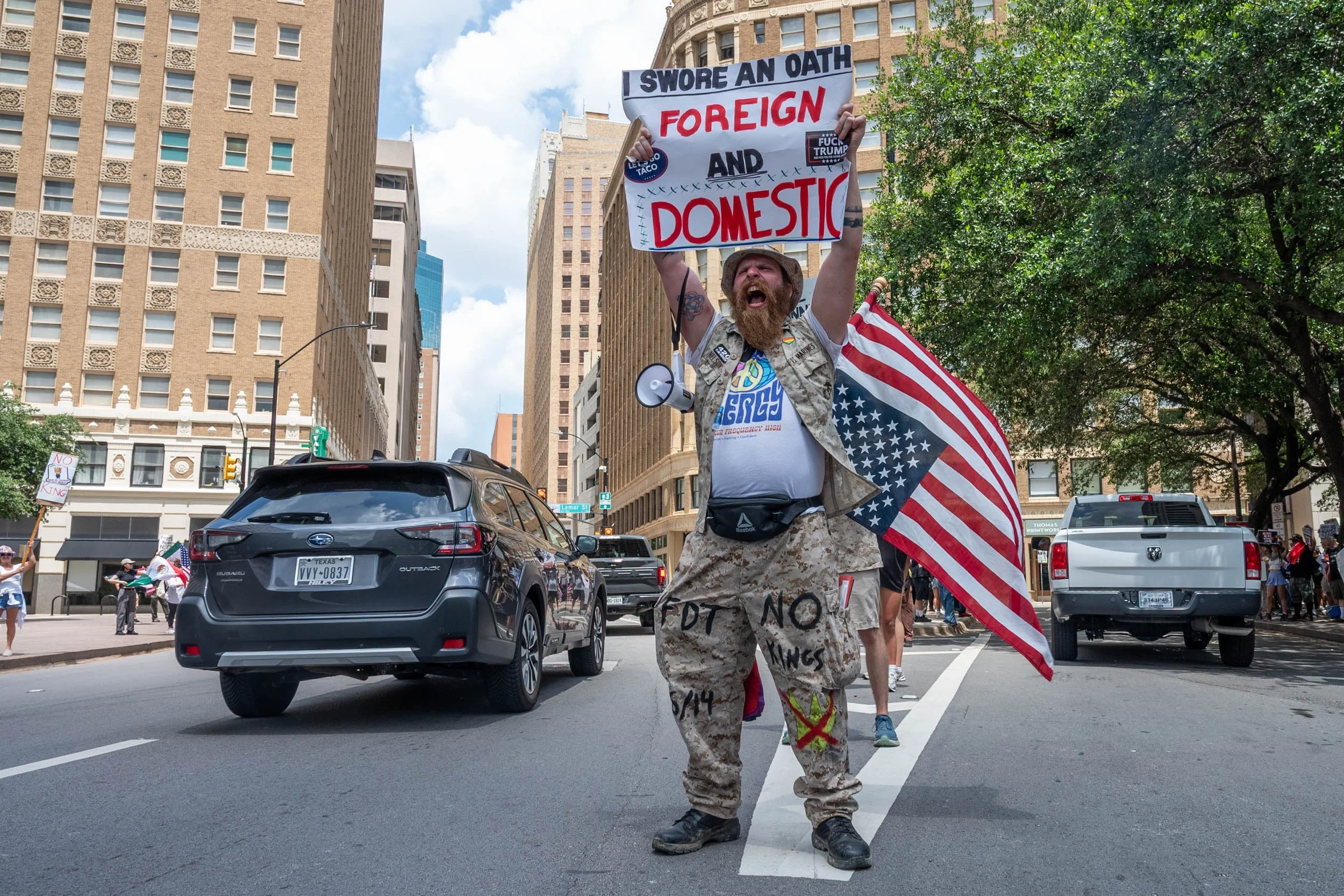 A man with a beard protesting in an urban area, holding a sign that says "I swore an oath foreign and domestic". He is wearing camouflage pants, a camouflage vest, and a hat, and has a flag draped over his shoulders. There are other protesters and ca