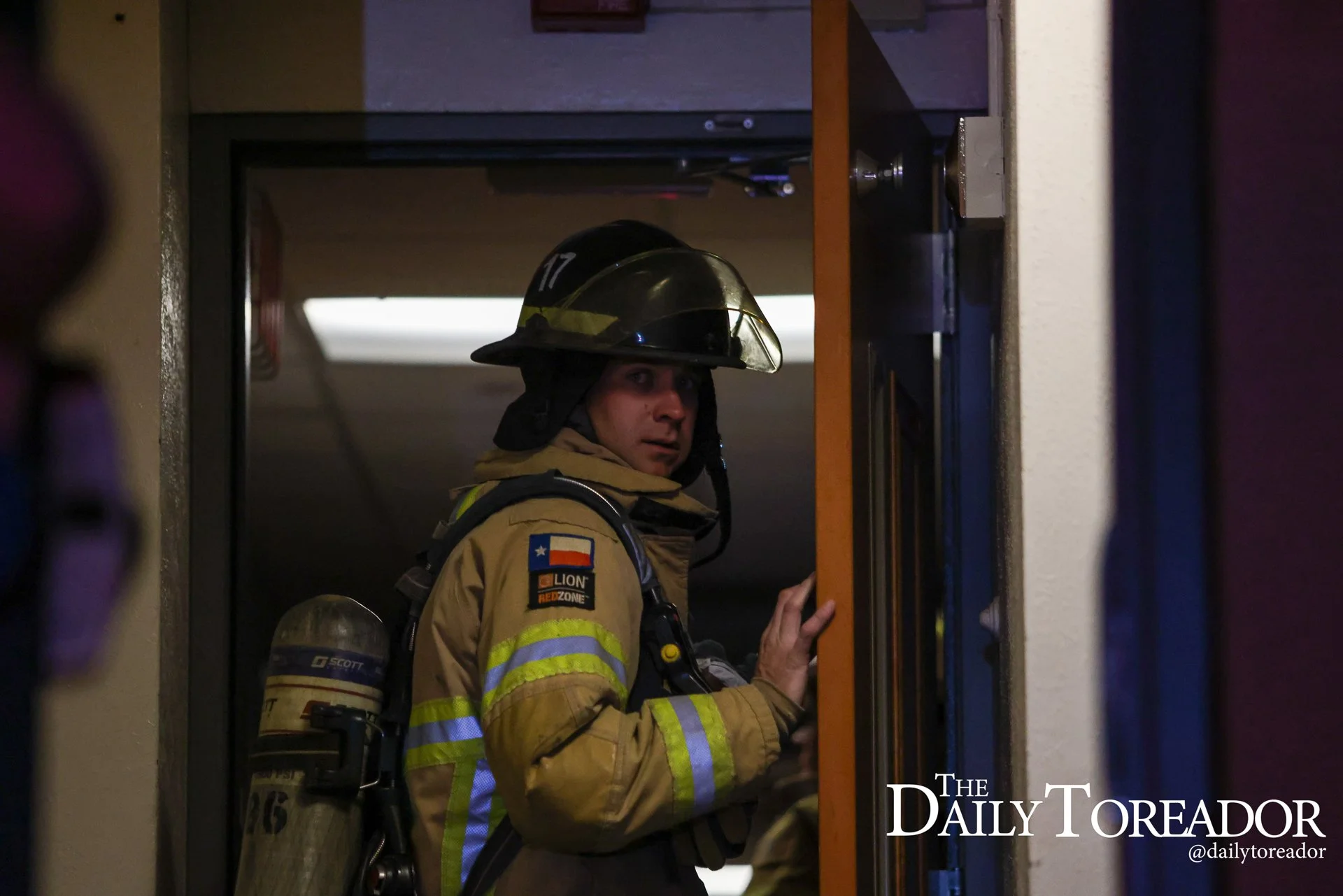 A firefighter in full gear, including a helmet with the number 17, stands in a doorway. The firefighter is looking over his shoulder, and the scene appears to be indoors, possibly after an emergency or fire. Part of a breathing apparatus is visible o