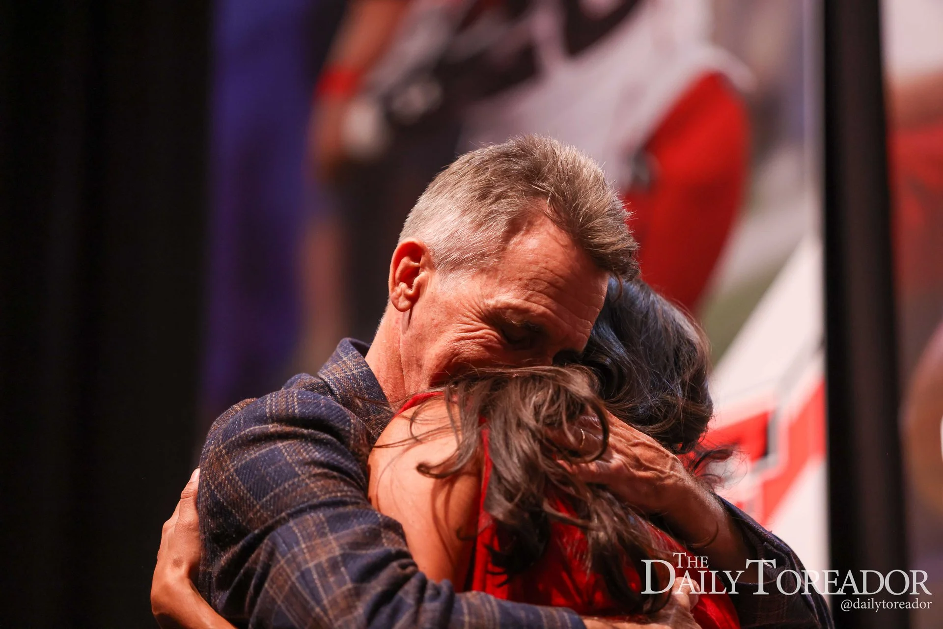 Two people hugging tightly, one with greying hair and the other with curly dark hair. They are embracing each other warmly in an indoor setting.