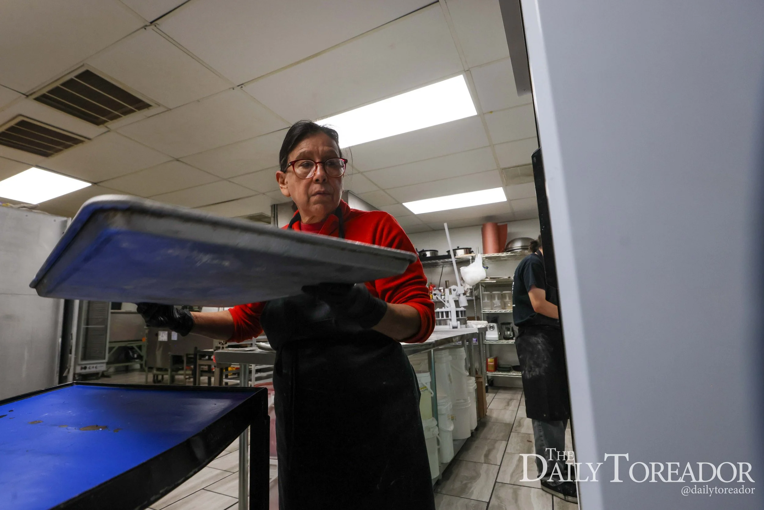 A woman in red shirt and glasses working in a commercial kitchen, standing behind stainless steel counters and shelves.