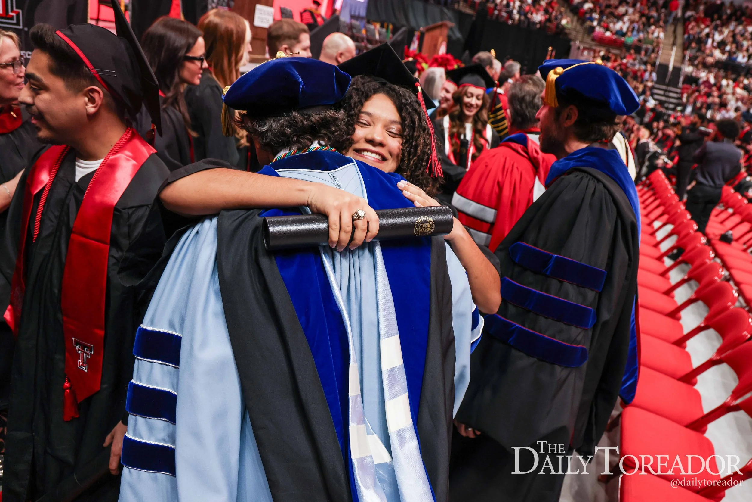 A group of graduates in caps and gowns, hugging at a graduation ceremony. The background shows more graduates and attendees.