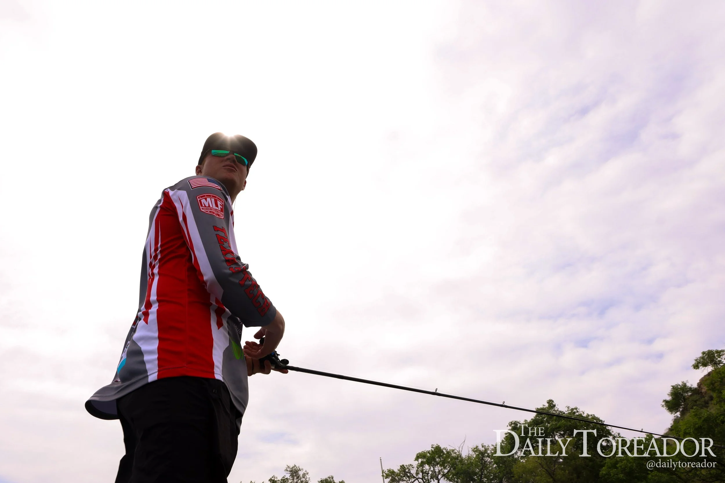 A man wearing sunglasses and a patterned sports jersey holding a fishing rod outdoors on a cloudy day with trees in the background.