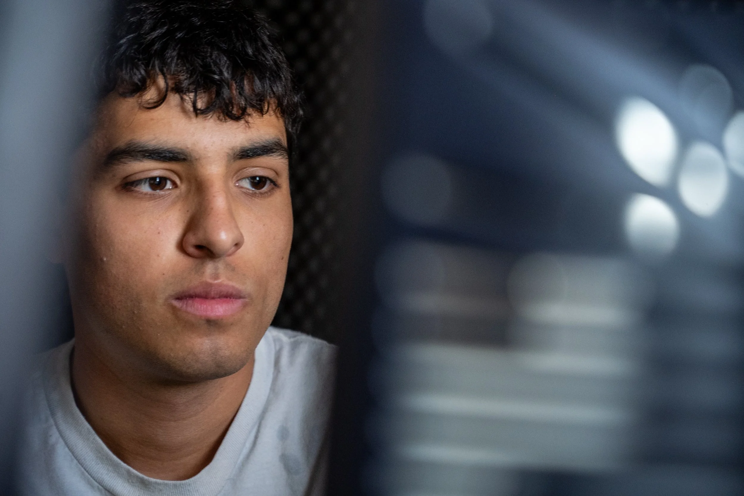 Close-up of a young man with curly dark hair, brown eyes, and light brown skin, looking thoughtfully to the side. He is wearing a light-colored shirt, and the background is blurred with circular light patterns.
