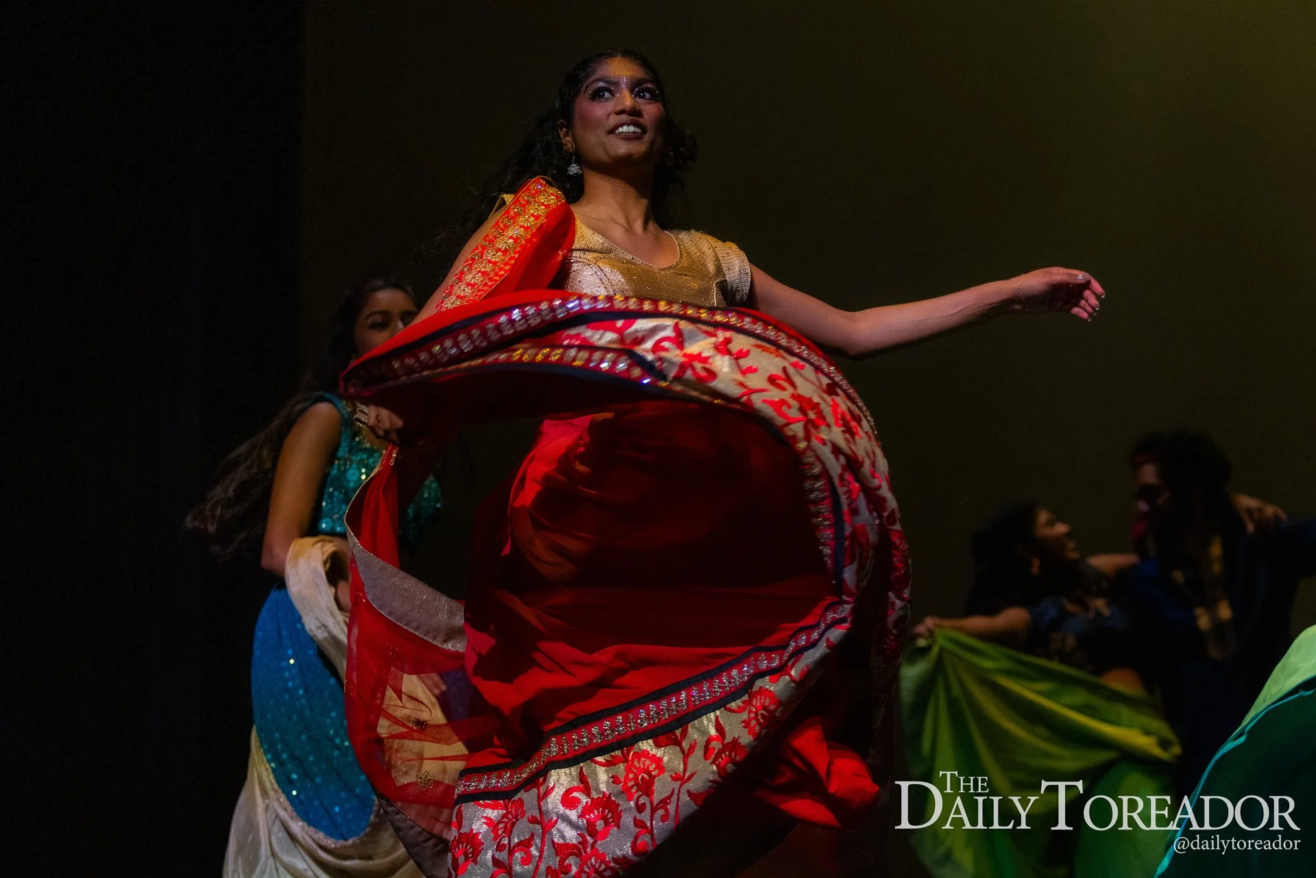 Woman in red and gold traditional dress dancing on stage with others in colorful outfits in the background at night.
