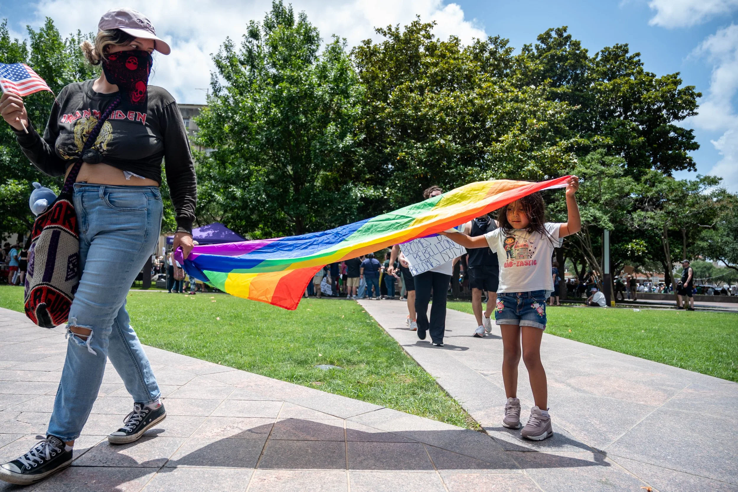 A diverse group of people participating in a parade or protest outdoors, carrying a rainbow flag and signs, with trees and a blue sky in the background.