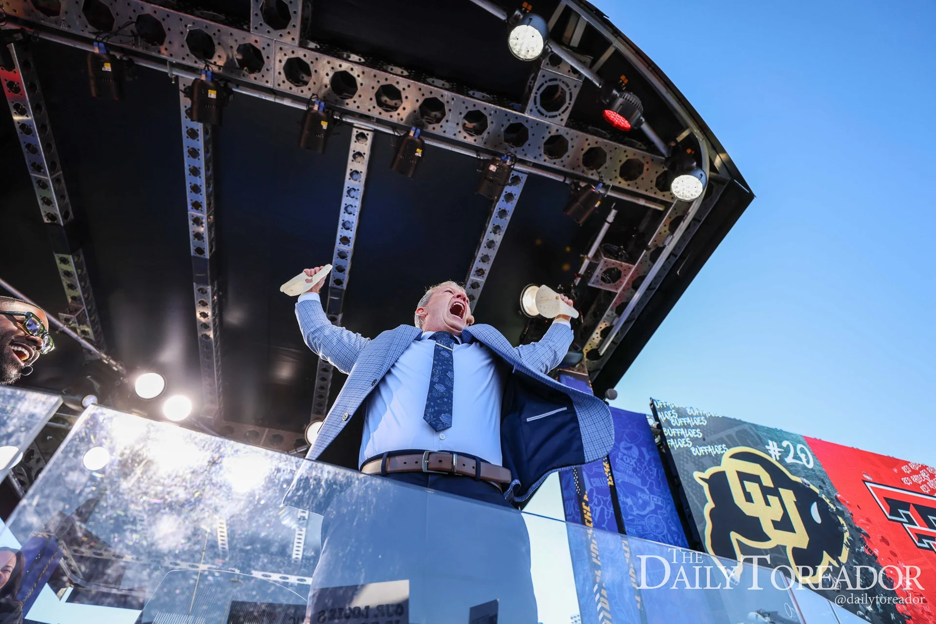A man in a blue checked blazer and tie cheering with his arms raised during a sports celebration on a stage with bright lights and a scoreboard in the background.