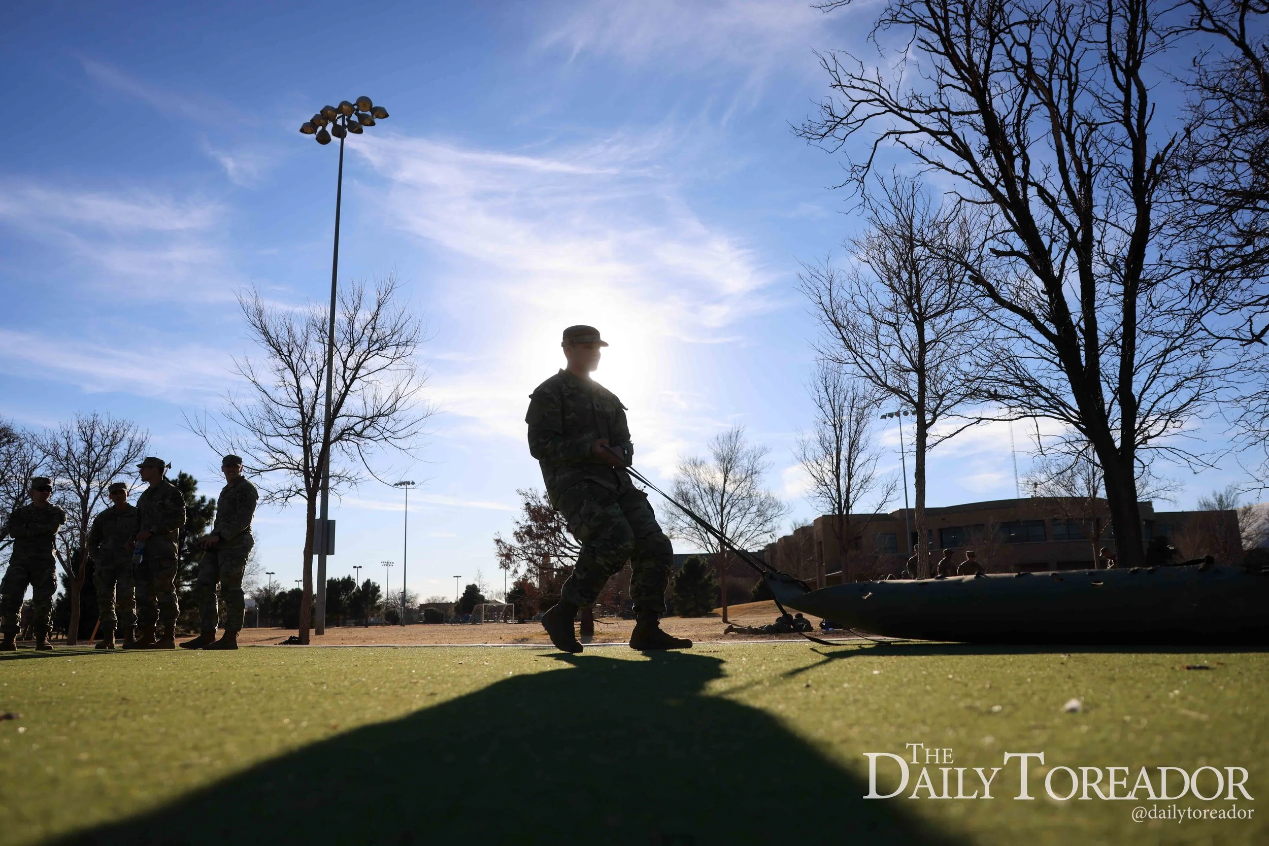 Military personnel gathered outdoors on a bright day with a clear blue sky, some standing in formation and one person in the foreground handling a kayak, with leafless trees and a building visible in the background.