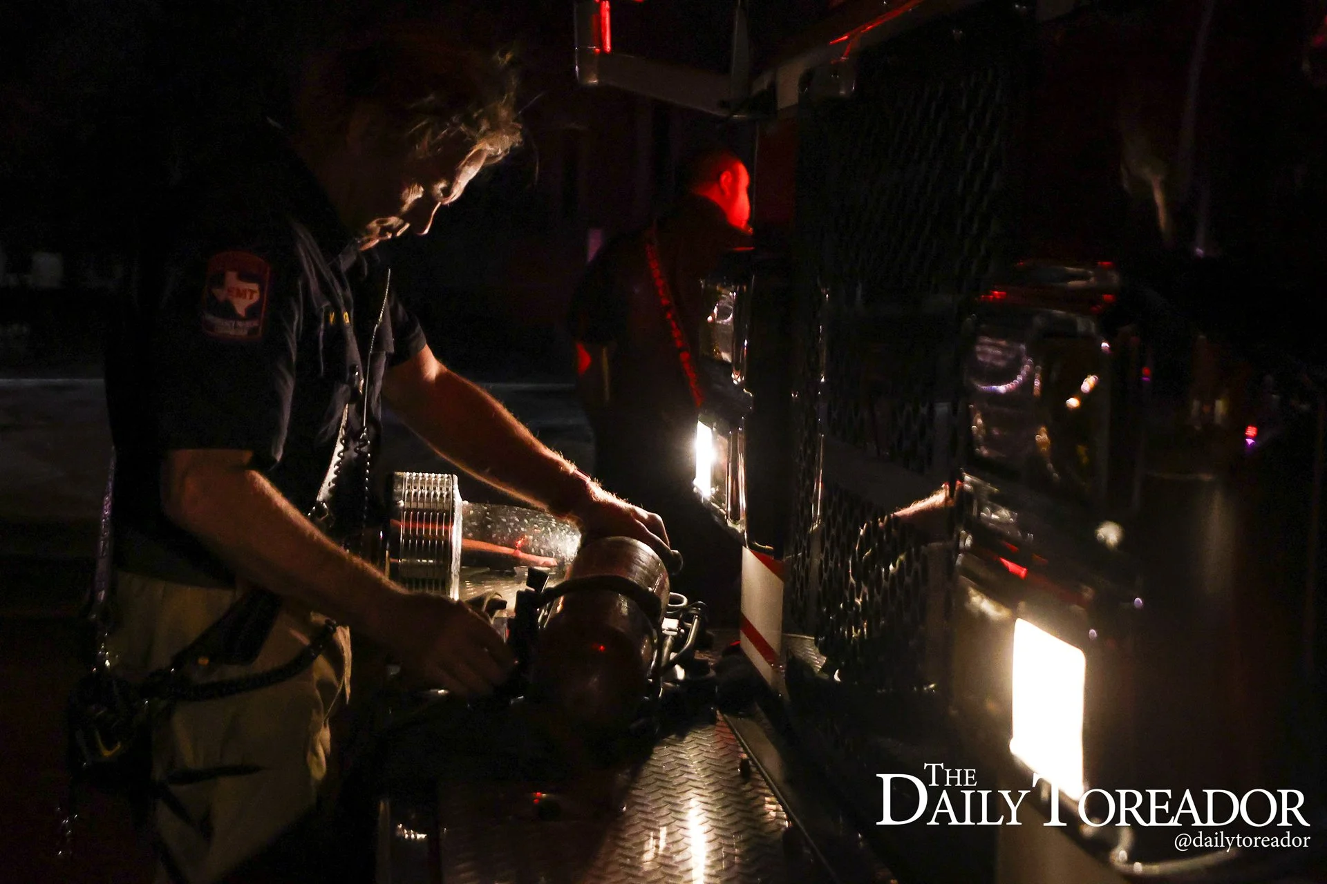 Nighttime scene of firefighter working near large emergency vehicle with reflective surfaces and equipment, illuminated by minimal lighting from the vehicle.
