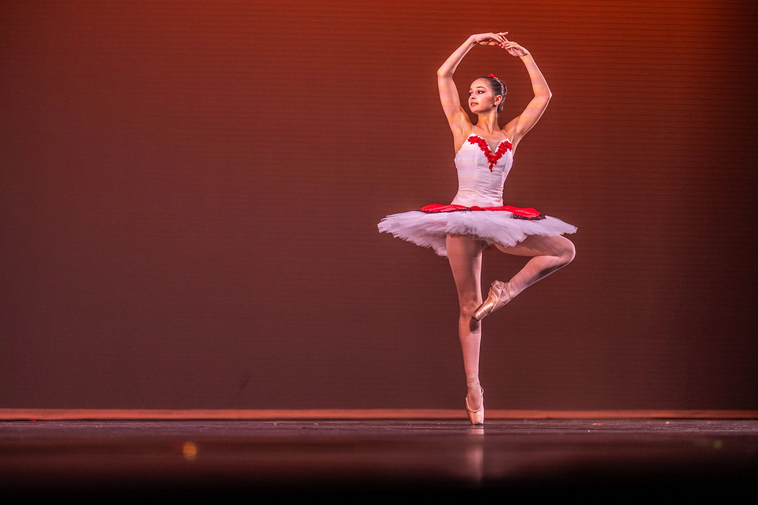 A ballet dancer in a white and red tutu performing on stage, balancing on one leg with arms curved above her head against a reddish background.