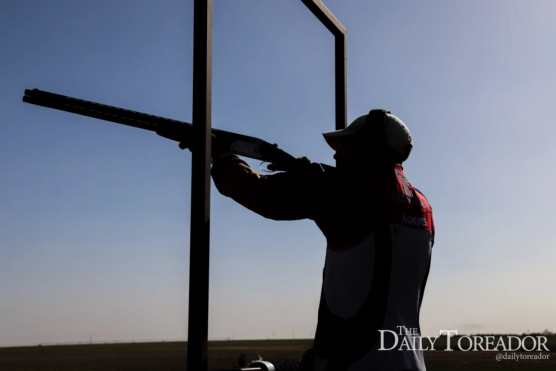 Silhouette of a person wearing a helmet and jacket, aiming a shotgun upwards through a metal frame against a clear sky and open landscape.