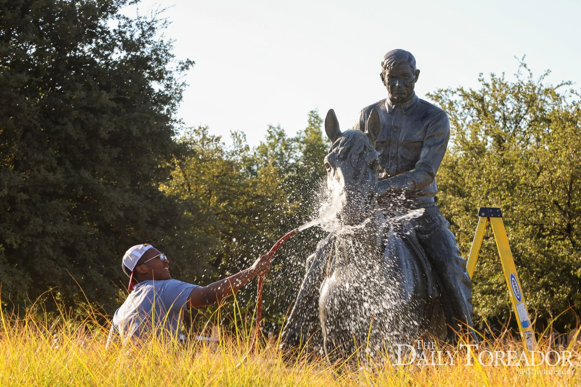 A young boy in a gray T-shirt and a white and gray cap spray water from a garden hose onto a bronze statue of a boy riding a horse in a park during daytime. The statue is surrounded by tall grass, with trees and a clear sky in the background.