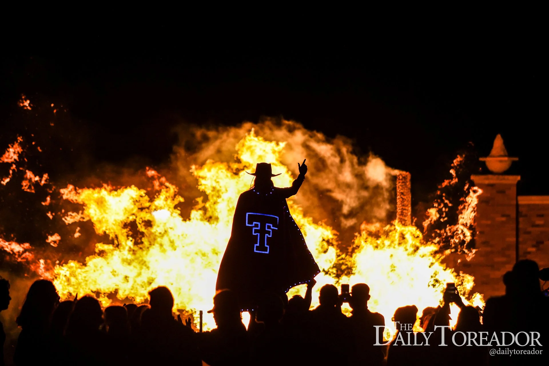 A person wearing a cowboy hat and a long cloak with the Tuff Toads logo stands amidst a large fire at night, with an audience watching. The sky is dark, and the scene is intense and dramatic.