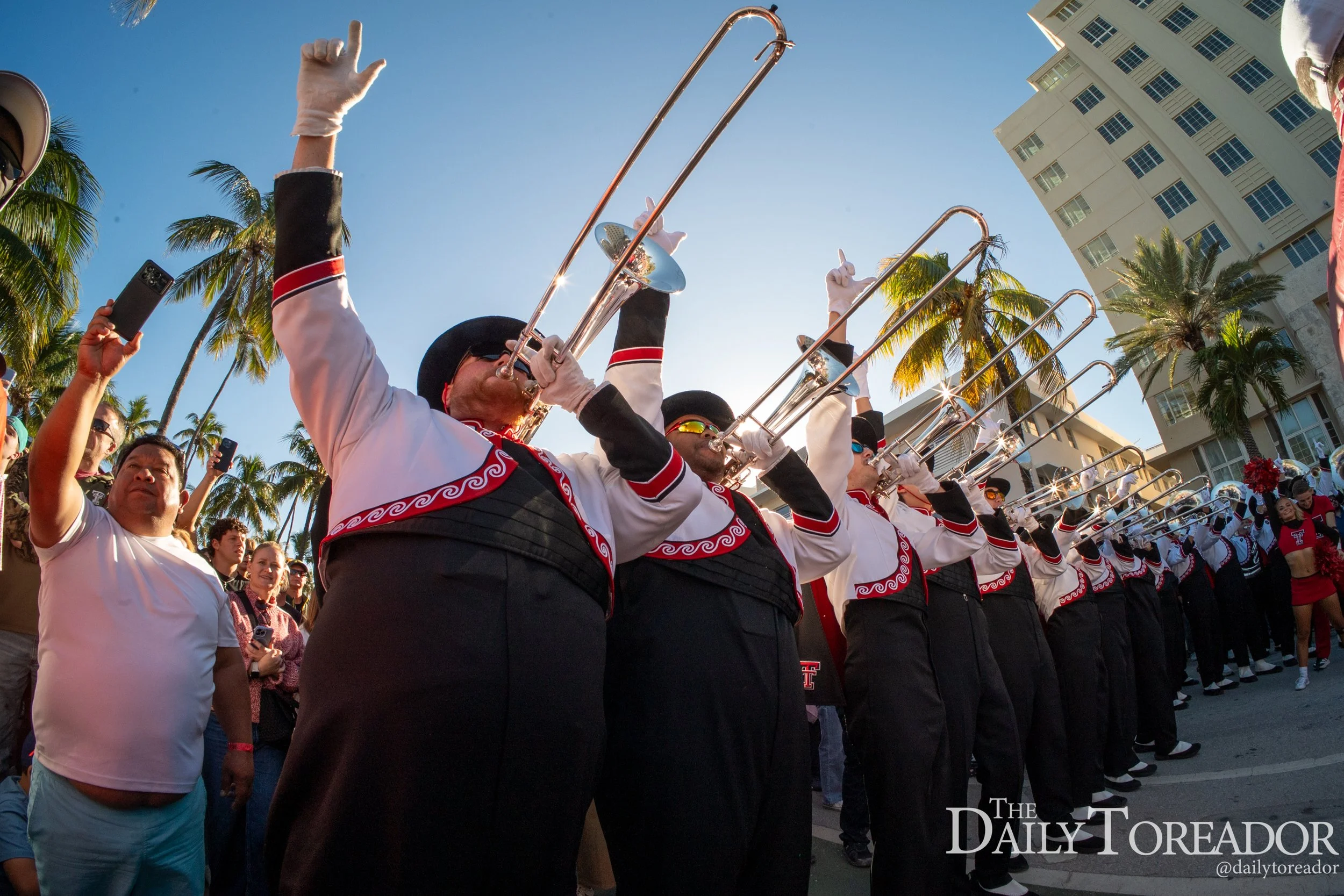 A parade with musicians playing trombones and other instruments, dressed in black and white uniforms with red accents, lined up in formation on a city street with tall buildings and palm trees in the background.