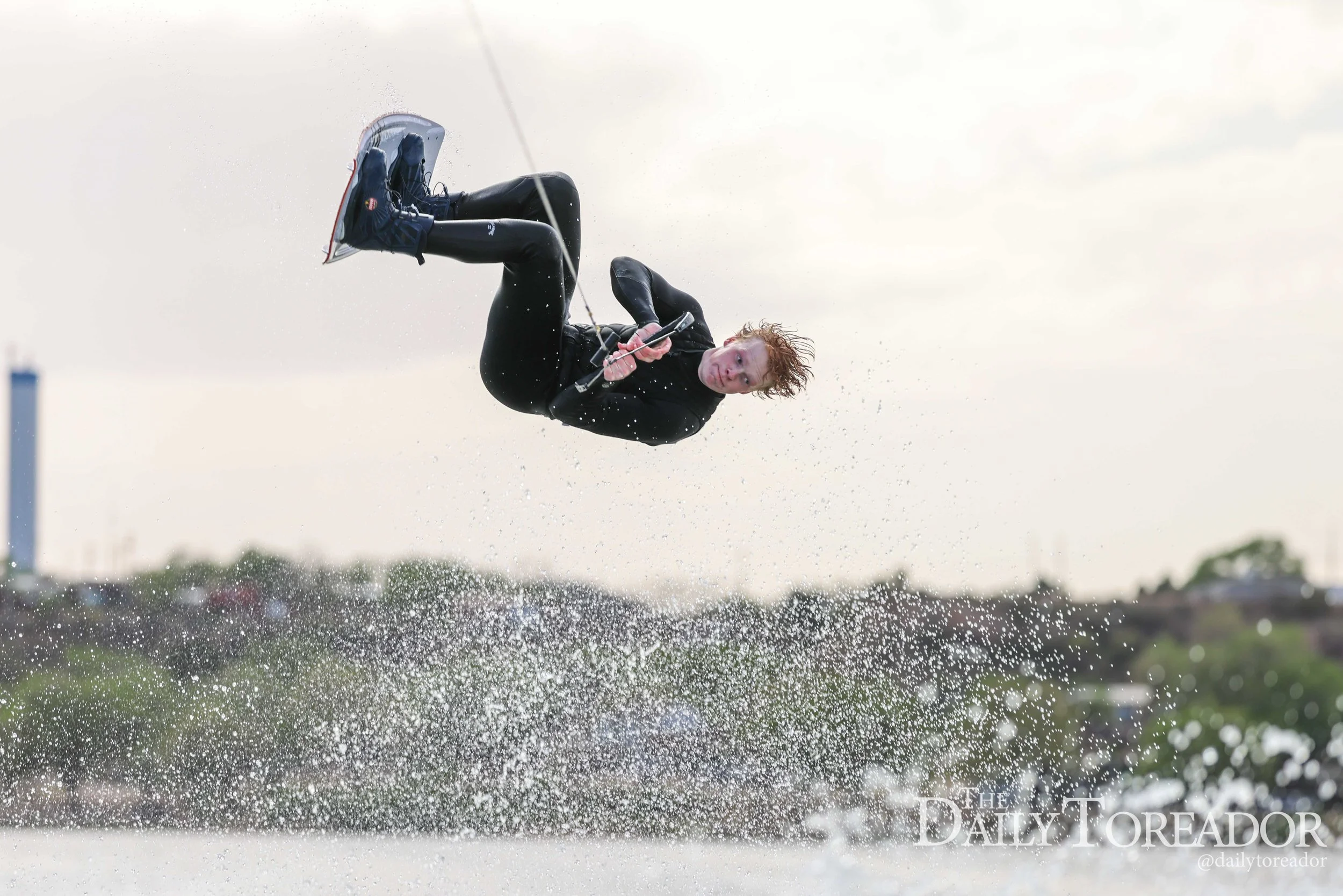Person wakeboarding on water, mid-air, with water splashing around, overcast sky.