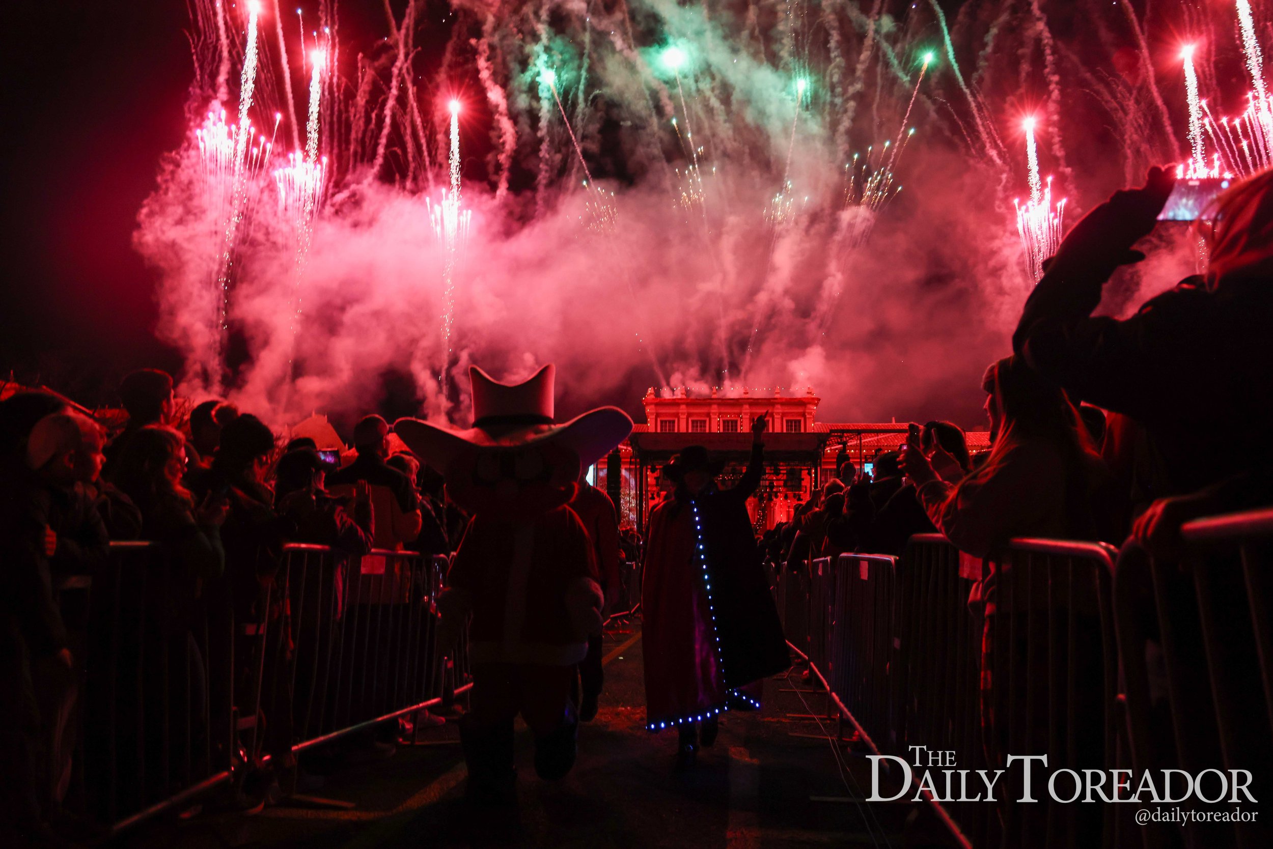 Crowd watching fireworks display at night, with colorful red and green explosions lighting up the sky, some people wearing hats, and barriers in the foreground.