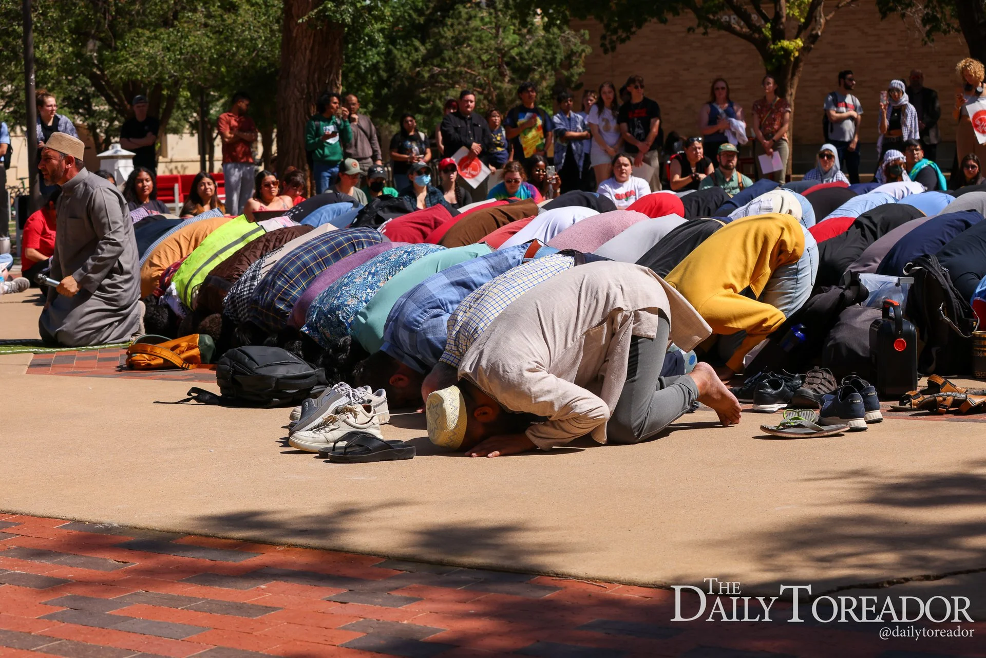 People praying outdoors with some kneeling and others standing, outdoors in a sunny area with trees and a brick building in the background.