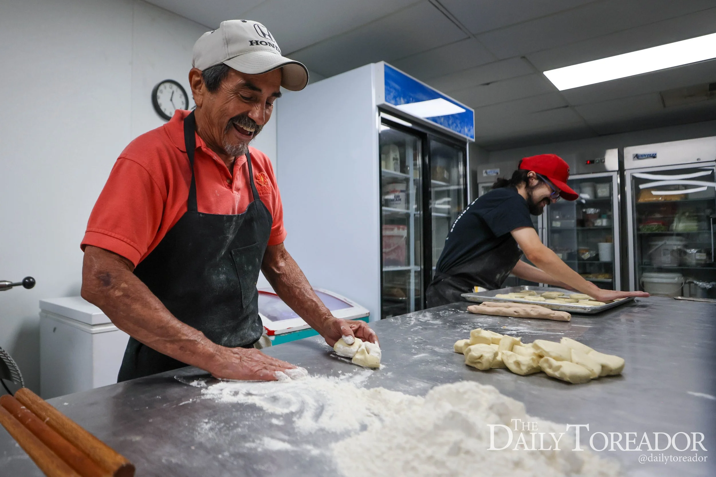 Two men preparing dough in a commercial kitchen, one older man with a mustache and beard wearing a baseball cap, and a younger man with long hair and glasses wearing a red cap, both working at a stainless steel table with dough pieces and flour.