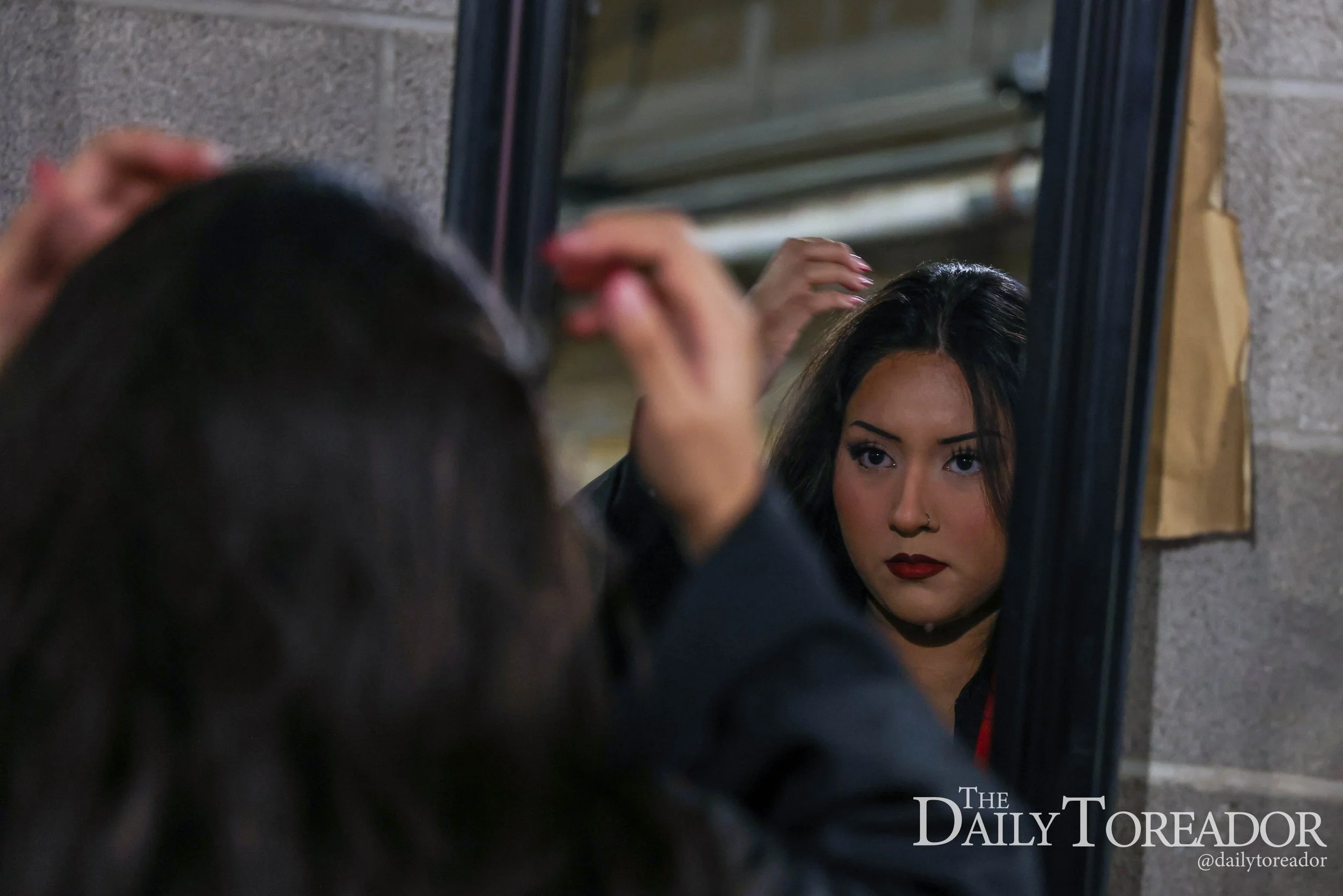A woman with dark hair and red lipstick looking into a mirror, adjusting her hair.