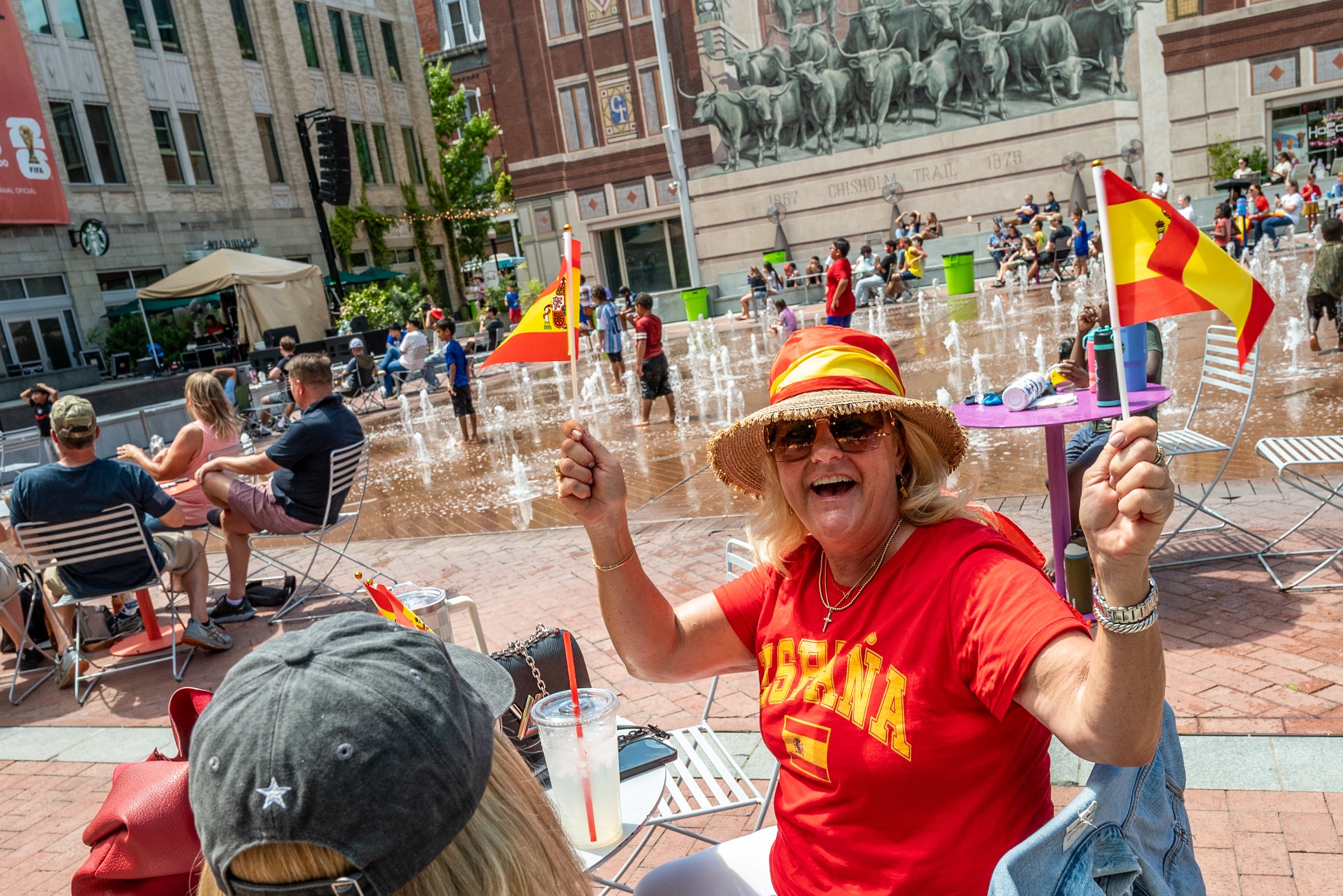 A woman wearing sunglasses, a straw hat, and a red T-shirt with yellow text, smiling and holding a Spanish flag in a lively outdoor setting with people sitting and standing around a fountain, some wearing red and yellow clothing, and a large bronze m
