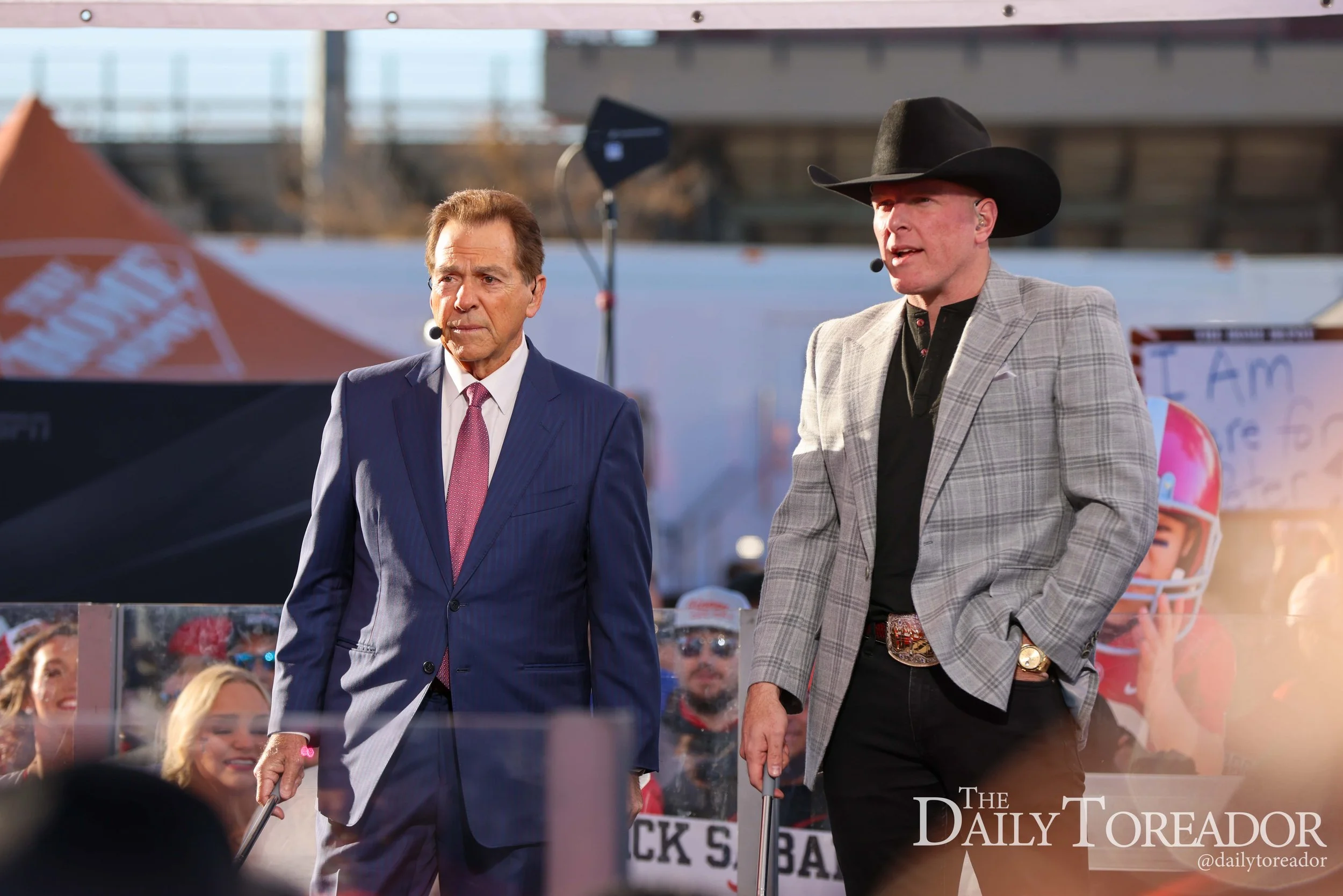 Two men wearing suits and cowboy hats walking in front of a crowd at an outdoor event, with signs and banners in the background.