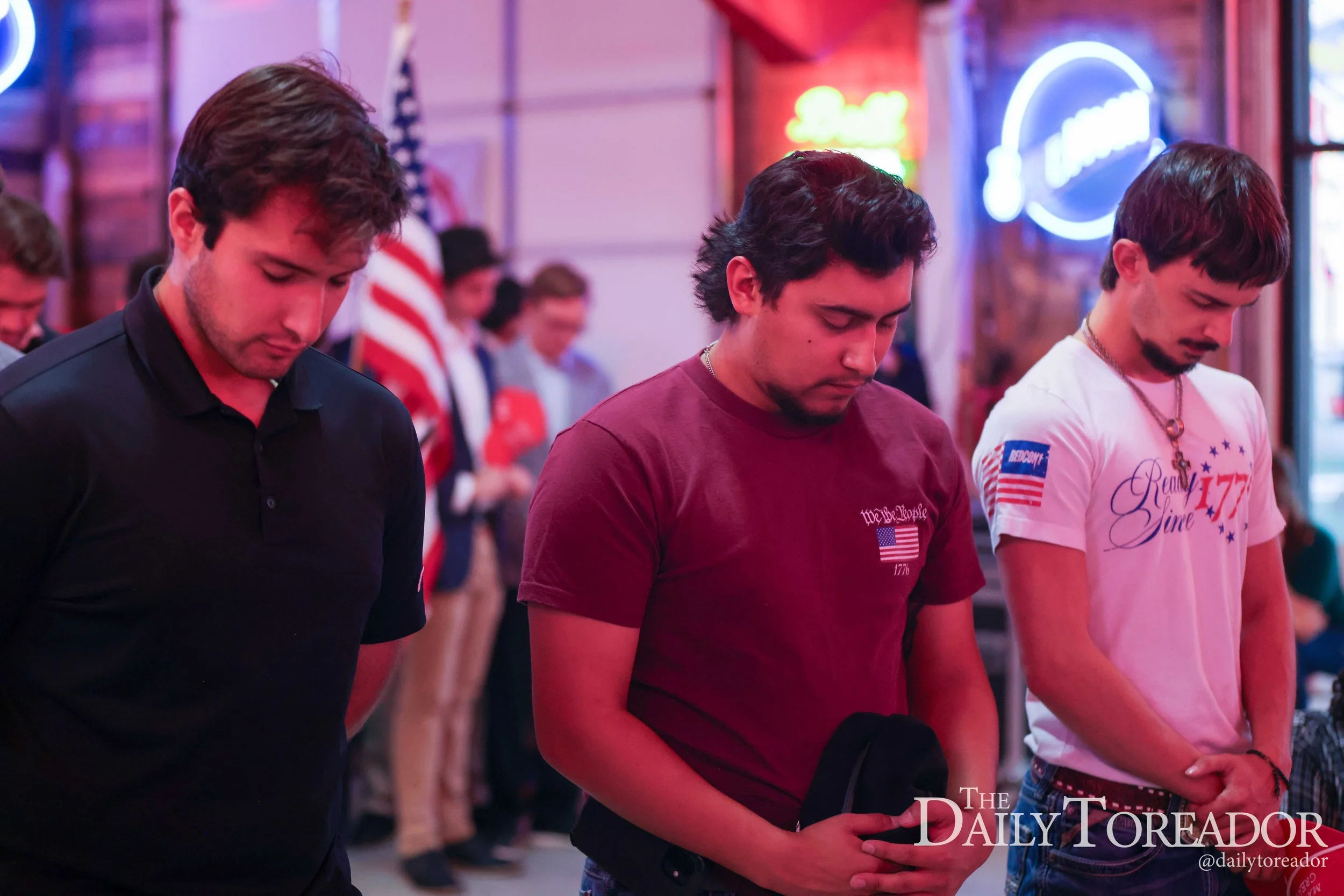 Three young men with closed eyes and bowed heads stand side by side in a room decorated with neon signs, with their hands held in front of them as if in prayer or reflection.