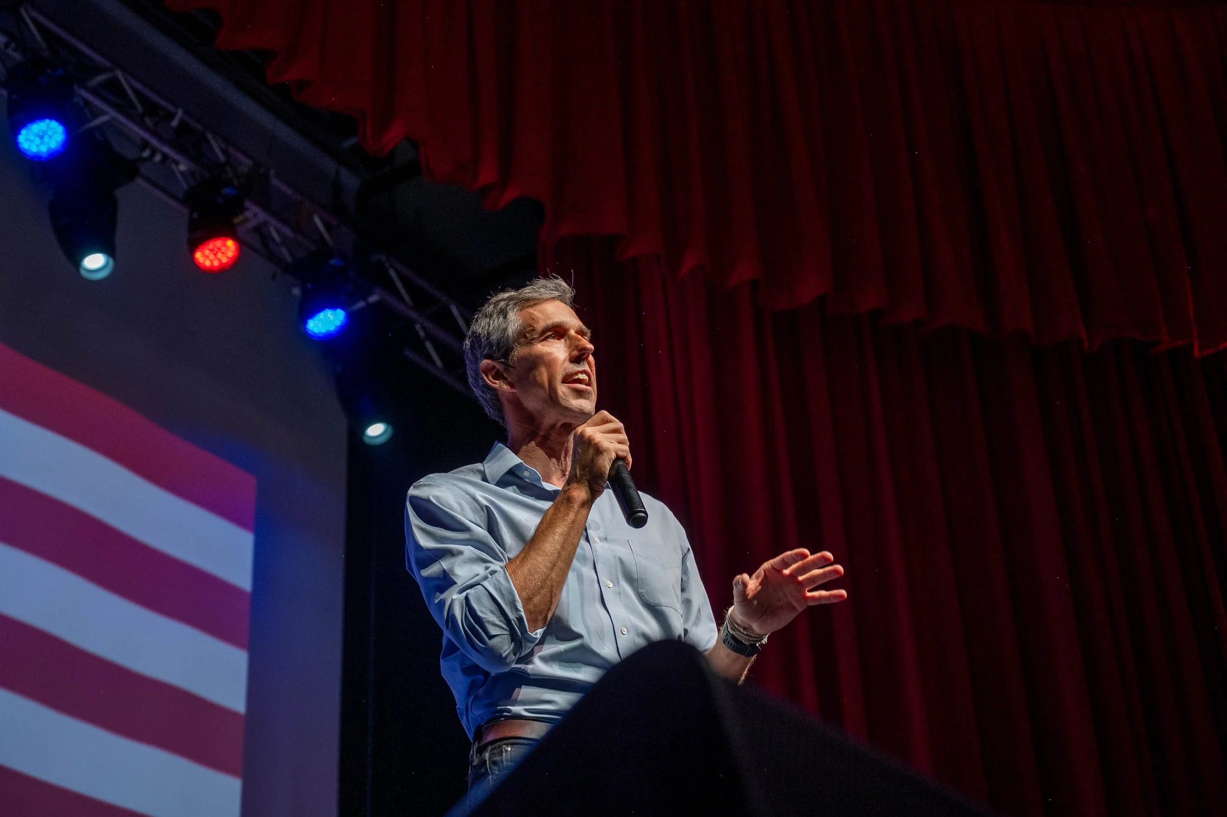 A man with gray hair and glasses, wearing a light blue shirt, speaking into a microphone during a presentation or speech on a stage with red curtains. Stage lights are visible above him.