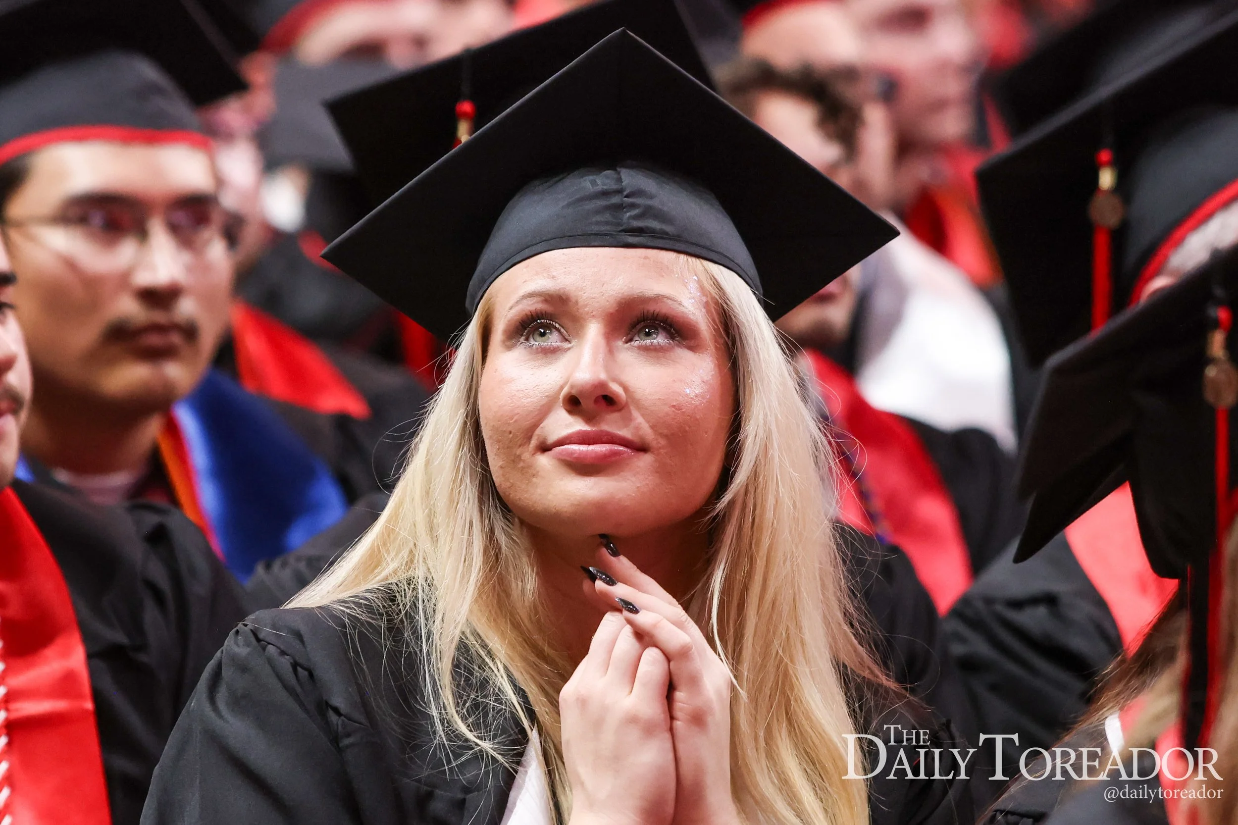 A blonde woman wearing a black graduation cap and gown, looking up with a thoughtful expression, surrounded by other graduates in similar attire.