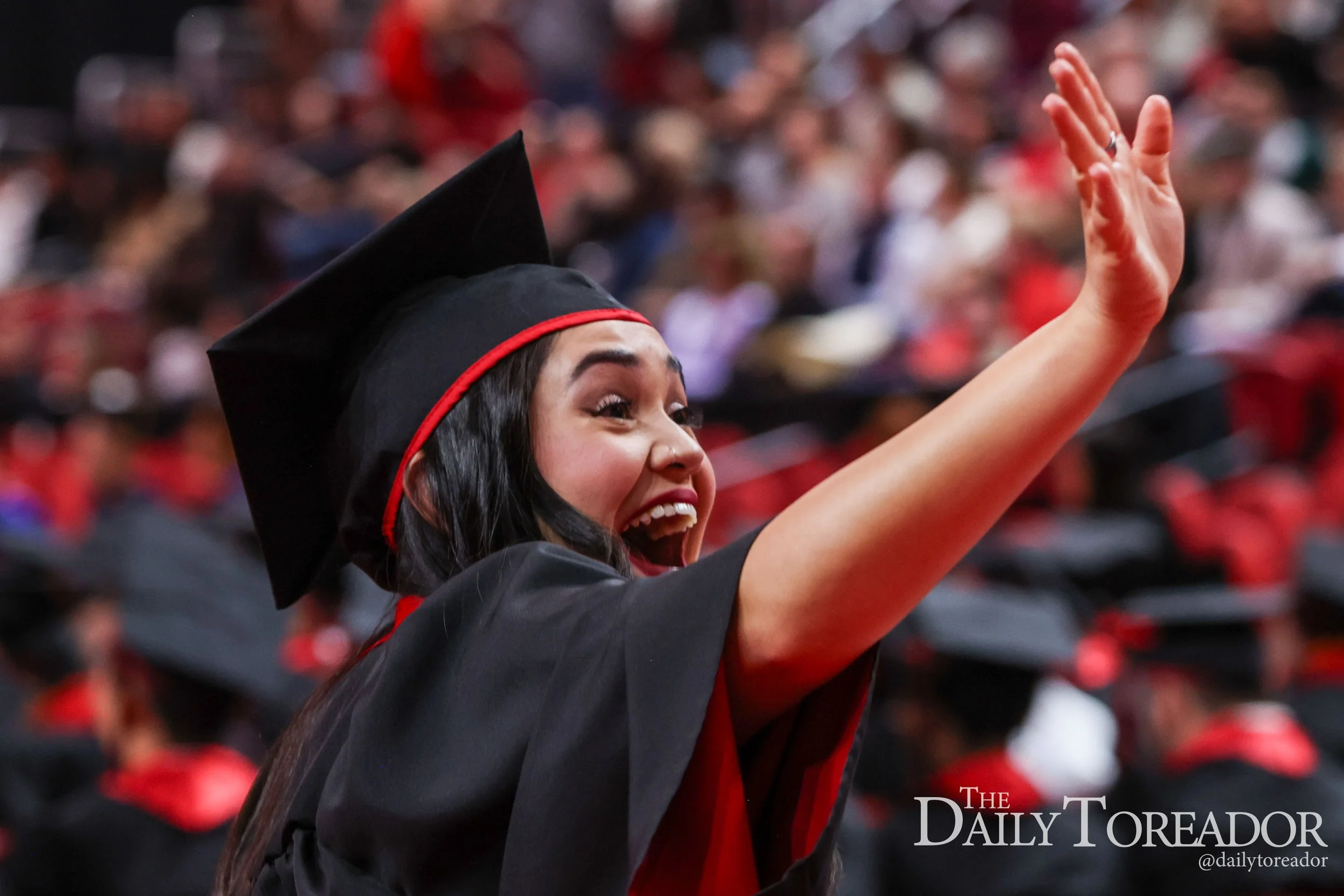 A young woman in a mortarboard cap and gown is celebrating at a graduation ceremony, raising her arm and smiling amid a crowd of graduates.