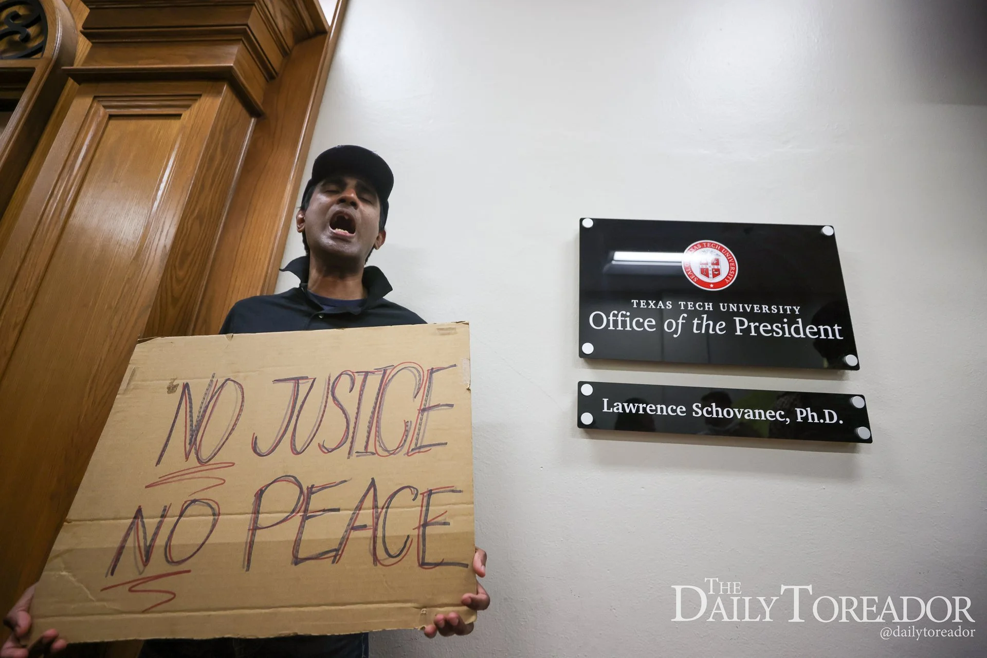 A person holding a cardboard sign that reads 'NO JUSTICE NO PEACE' in front of a wall with a plaque for Texas Tech University's Office of the President. The person is wearing a black shirt and a black cap, with their face partially visible.