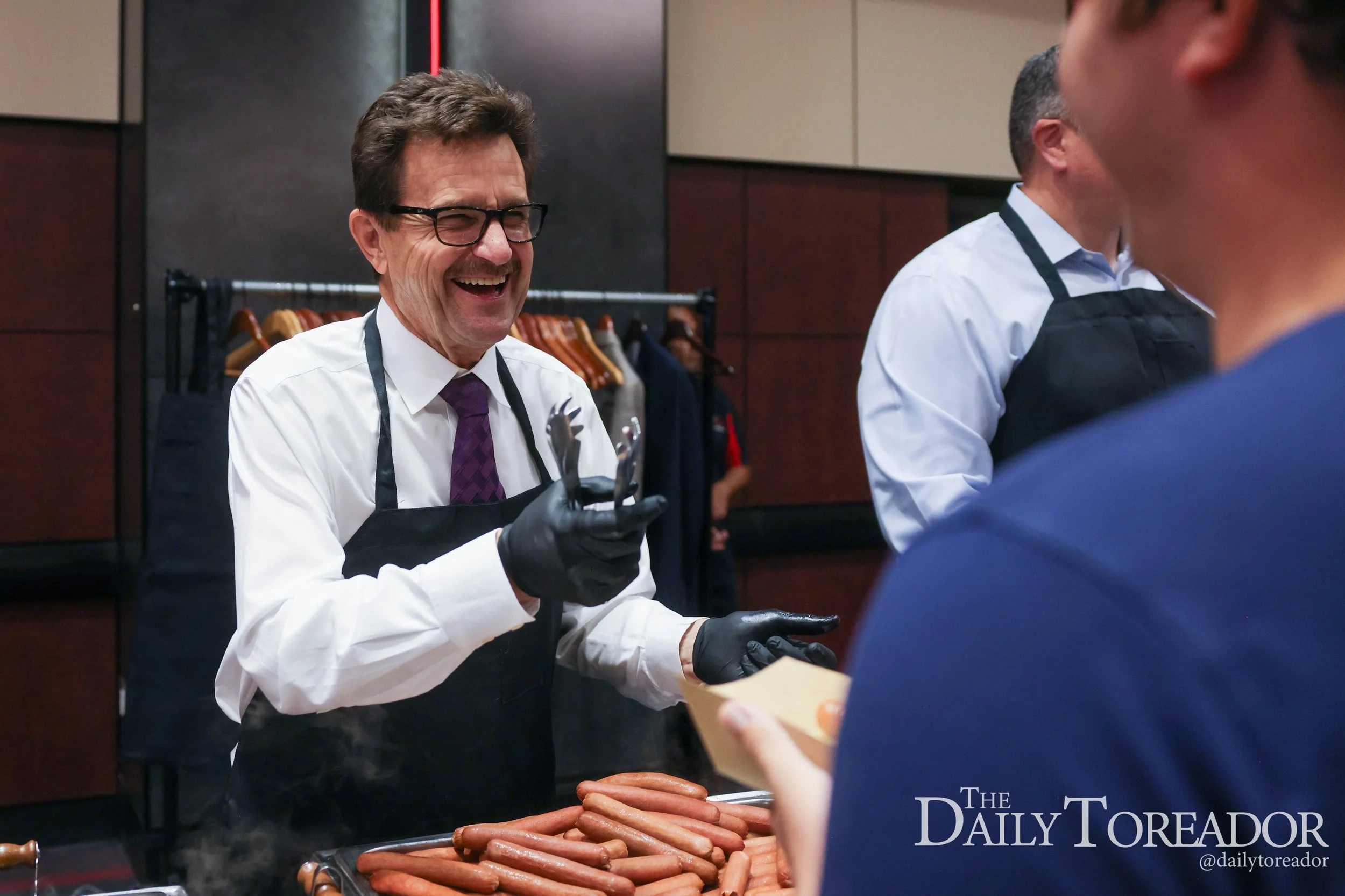 A man in glasses and a white shirt laughs while serving hot dogs at a food stand. There are other people in line, and the background shows a coat rack and dark-colored walls.