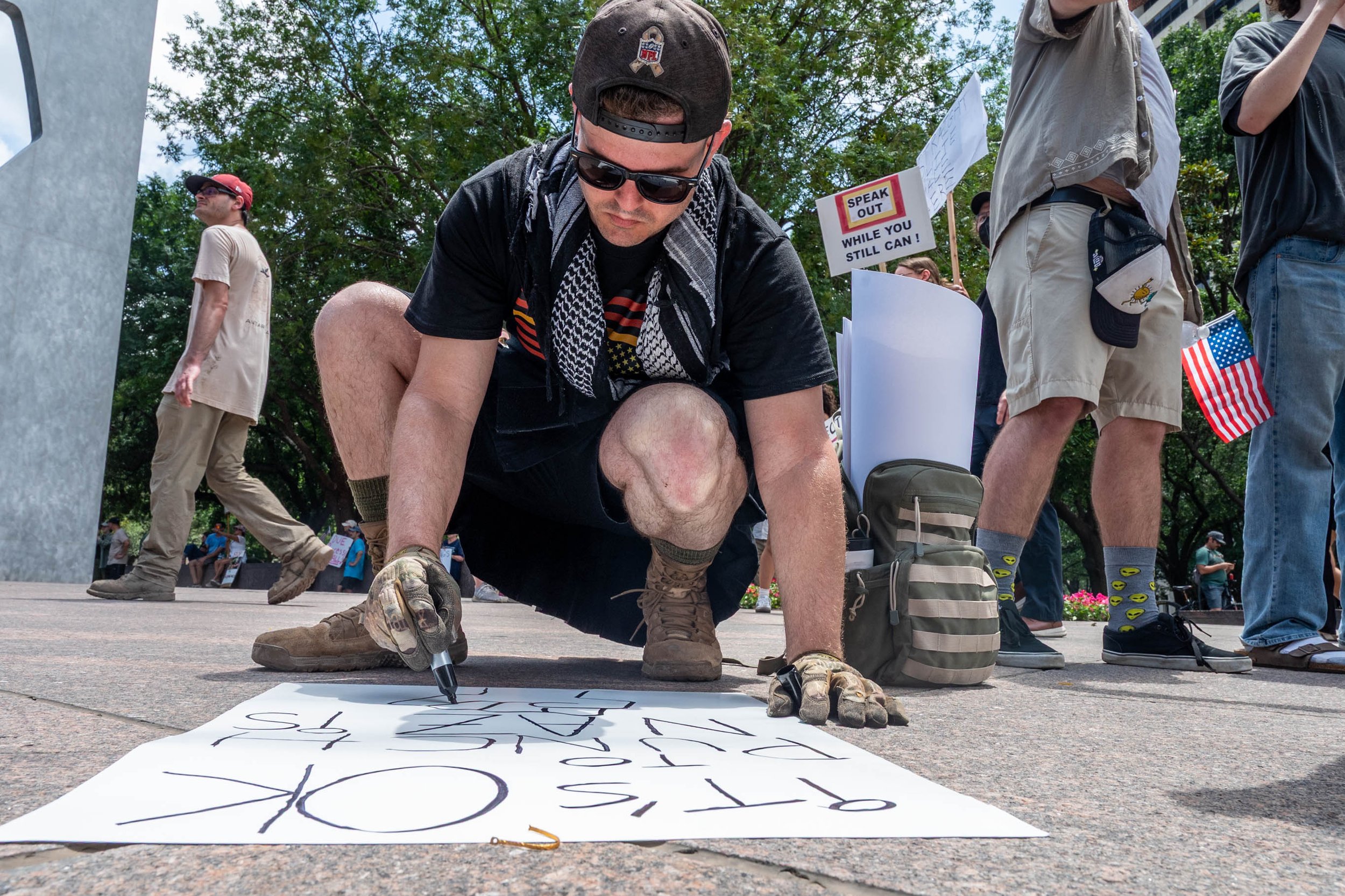 A man kneels on the ground signing a protest banner with a black marker at a protest event. Other protesters and signs are visible in the background, including a sign that says 'Speak out while you still can!'