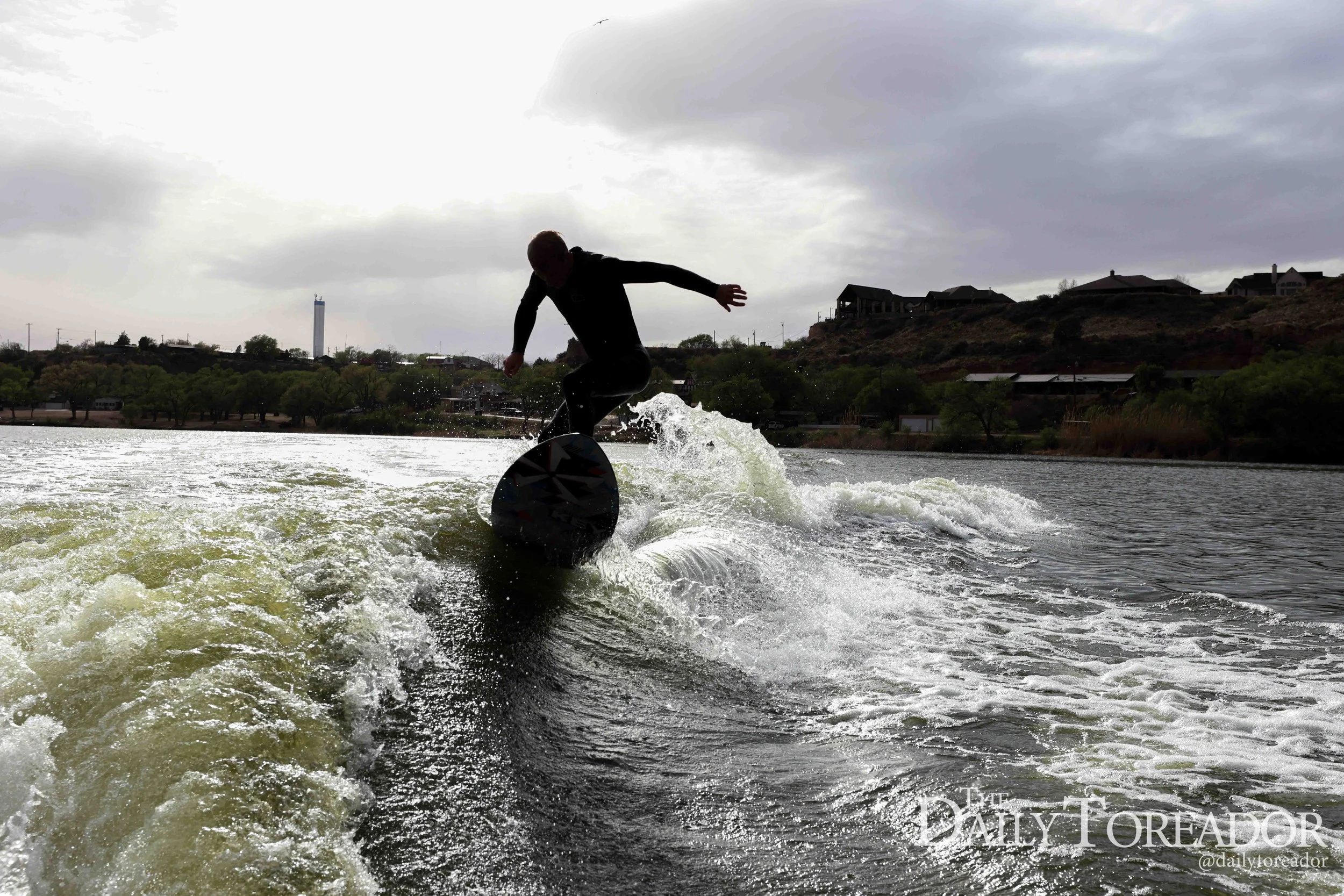 Silhouette of a person wakeboarding on a lake amid overcast weather, with houses and hills in the background.