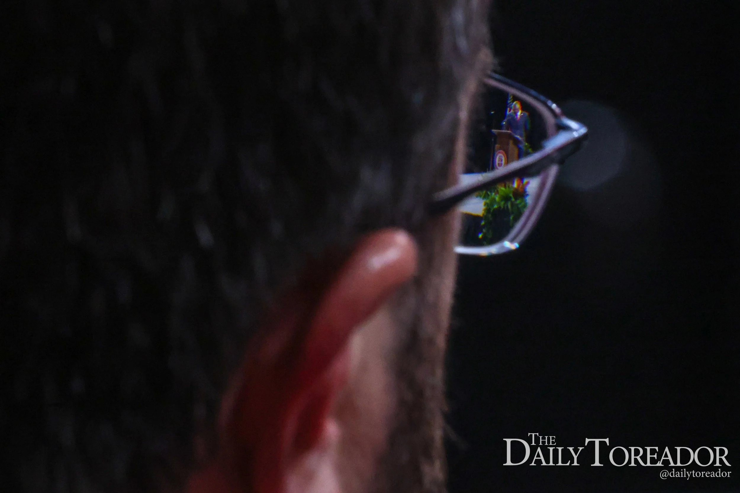 A person's face wearing glasses, seen from behind in a dark setting. The reflection in the glasses shows a woman standing near a green plant, possibly giving a presentation or speech.