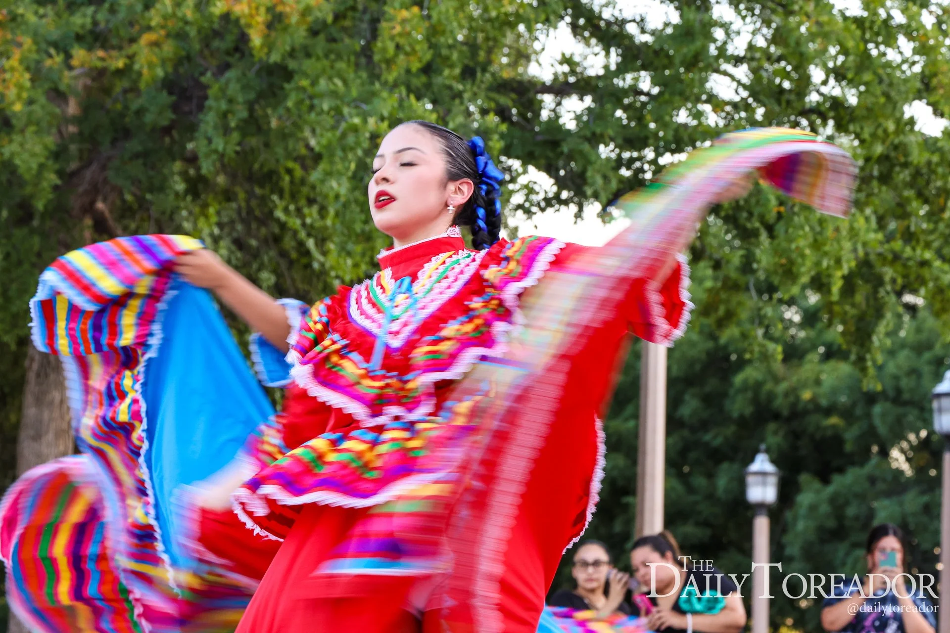 A woman in a vibrant red, rainbow-trimmed traditional dress dancing outdoors, with green trees in the background and onlookers watching.