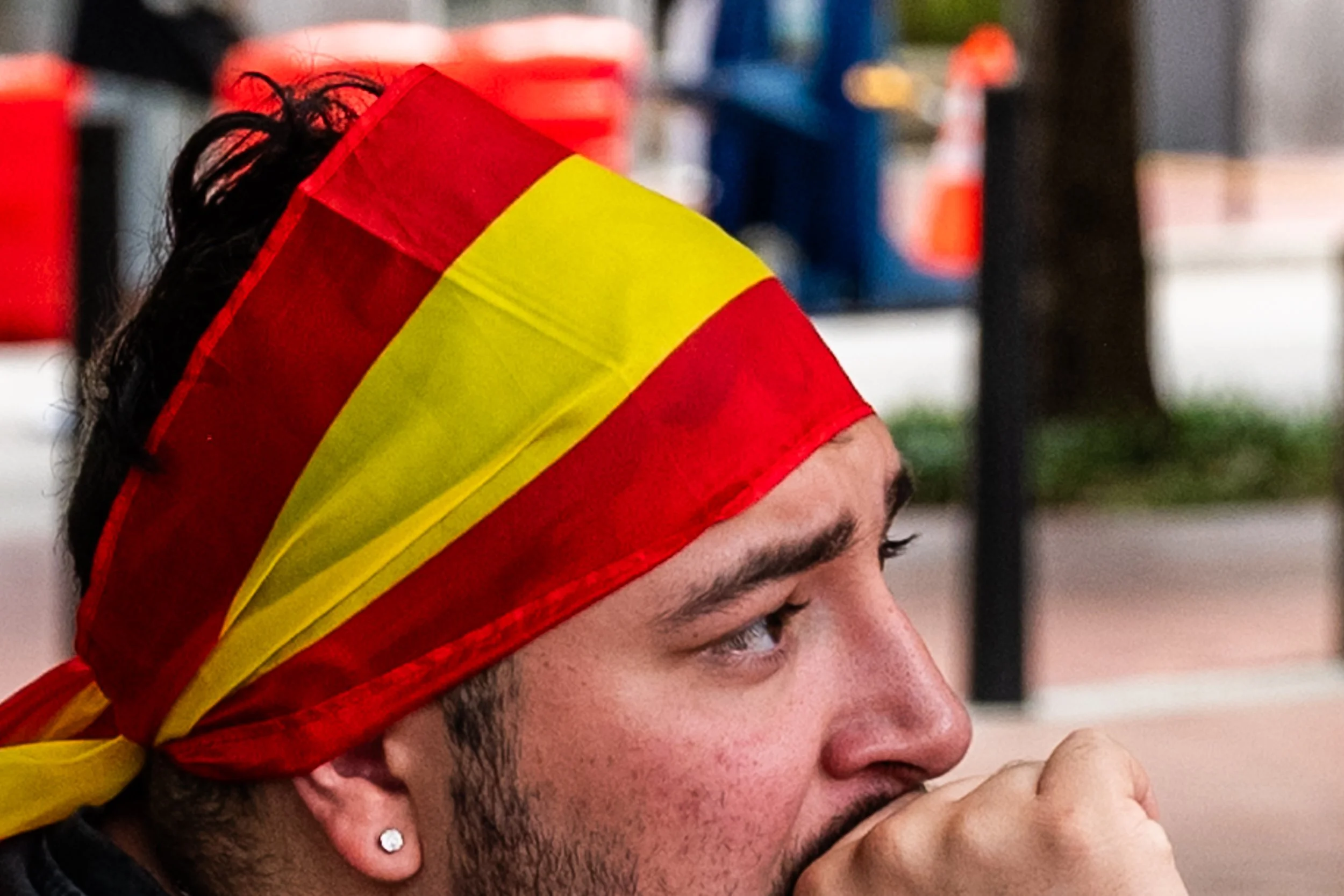 Close-up of a man wearing a red and yellow bandana, with dark hair and facial hair, touching his lips with his hand, with blurred background.