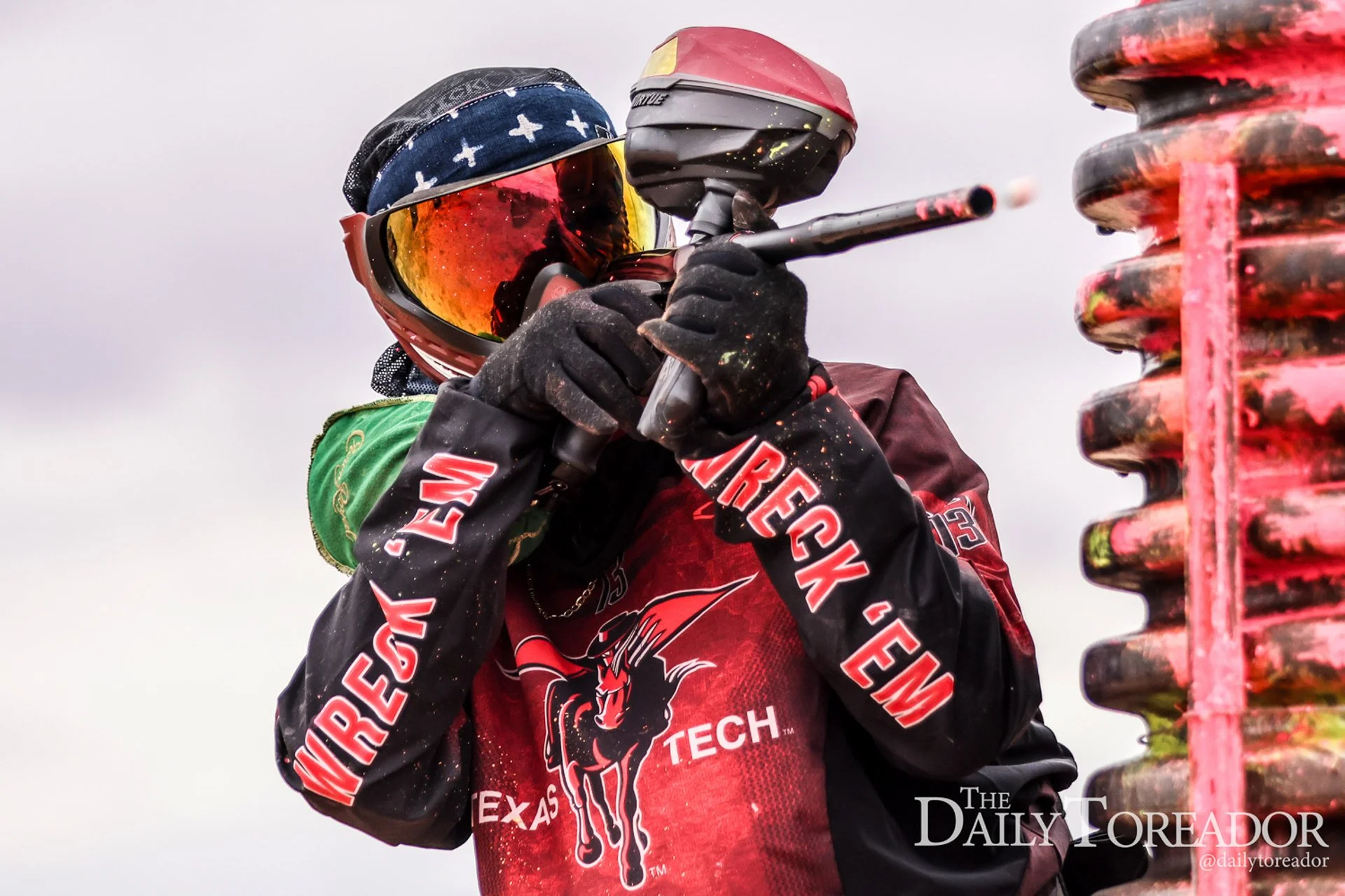 A person dressed in a Red and Black sports uniform holding a paintball marker with a red hopper, wearing reflective goggles and a bandana on their head, standing next to a stack of colorful paintball pods.