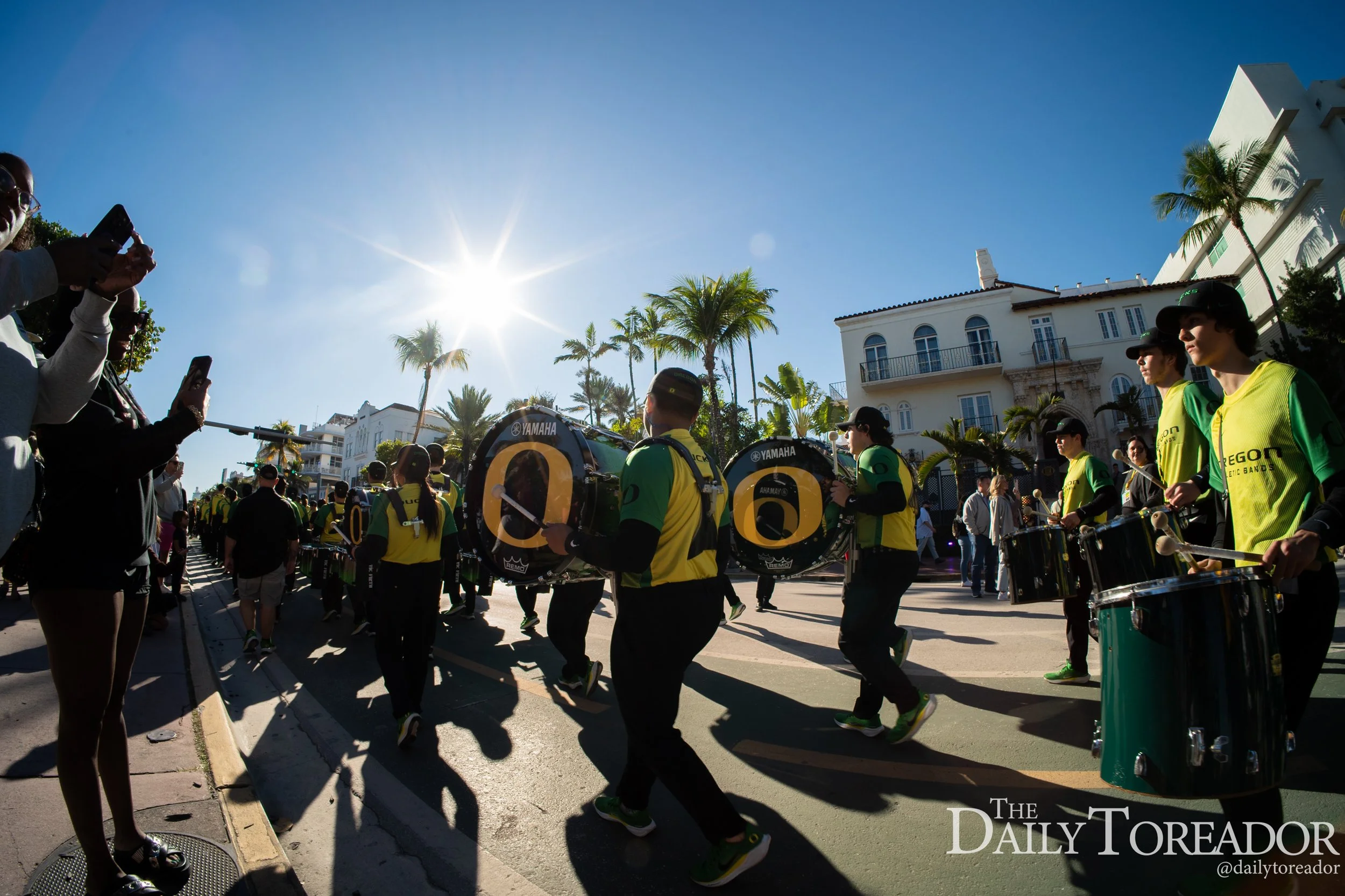 A parade with musicians in yellow and green uniforms playing drums on a sunny street lined with palm trees and white buildings.