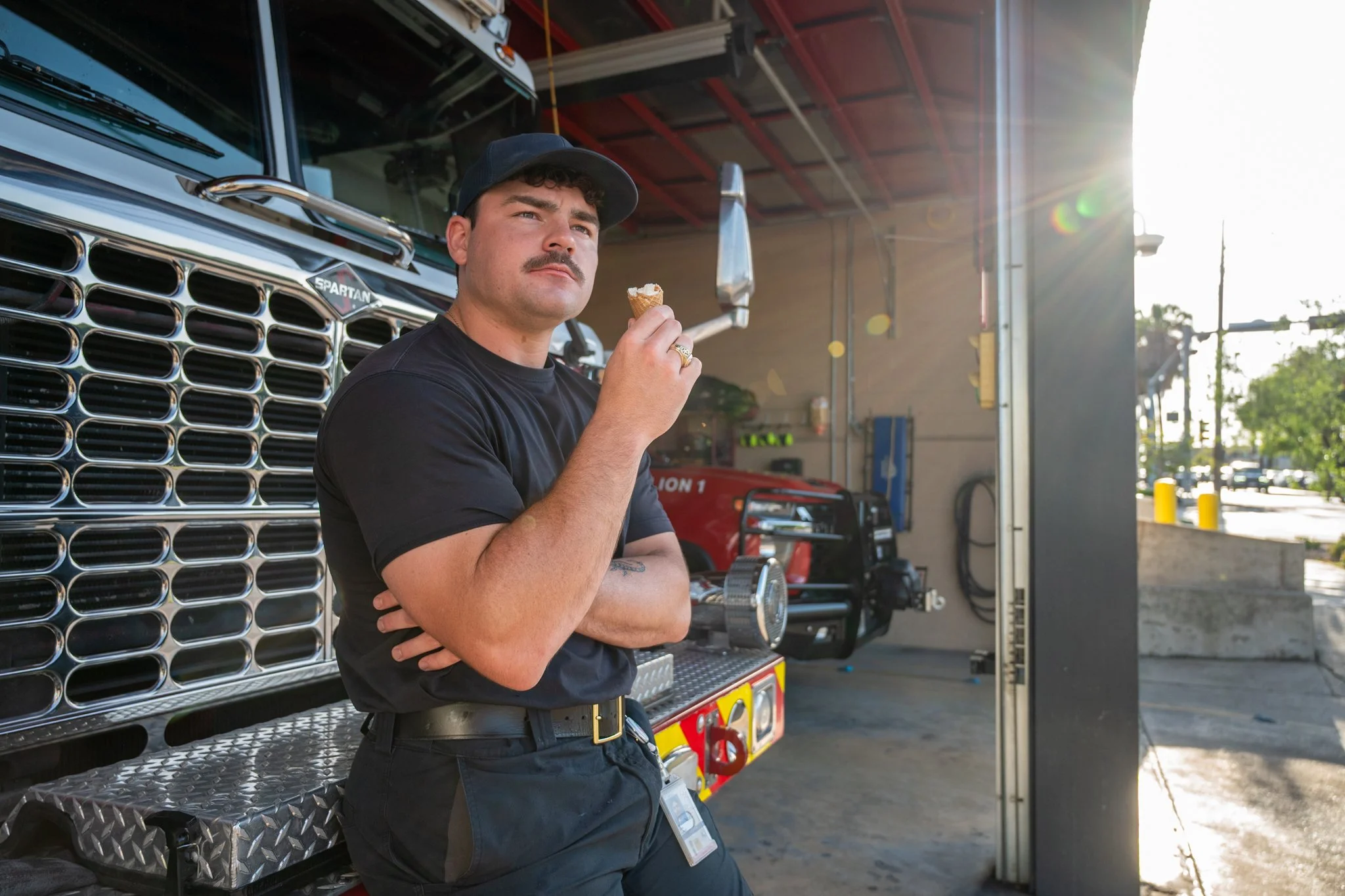 A young man with dark hair and a mustache, wearing a black cap and black T-shirt, stands beside a fire truck at a fire station, holding an ice cream cone and looking off into the distance with a thoughtful expression, sun shining from the right side 