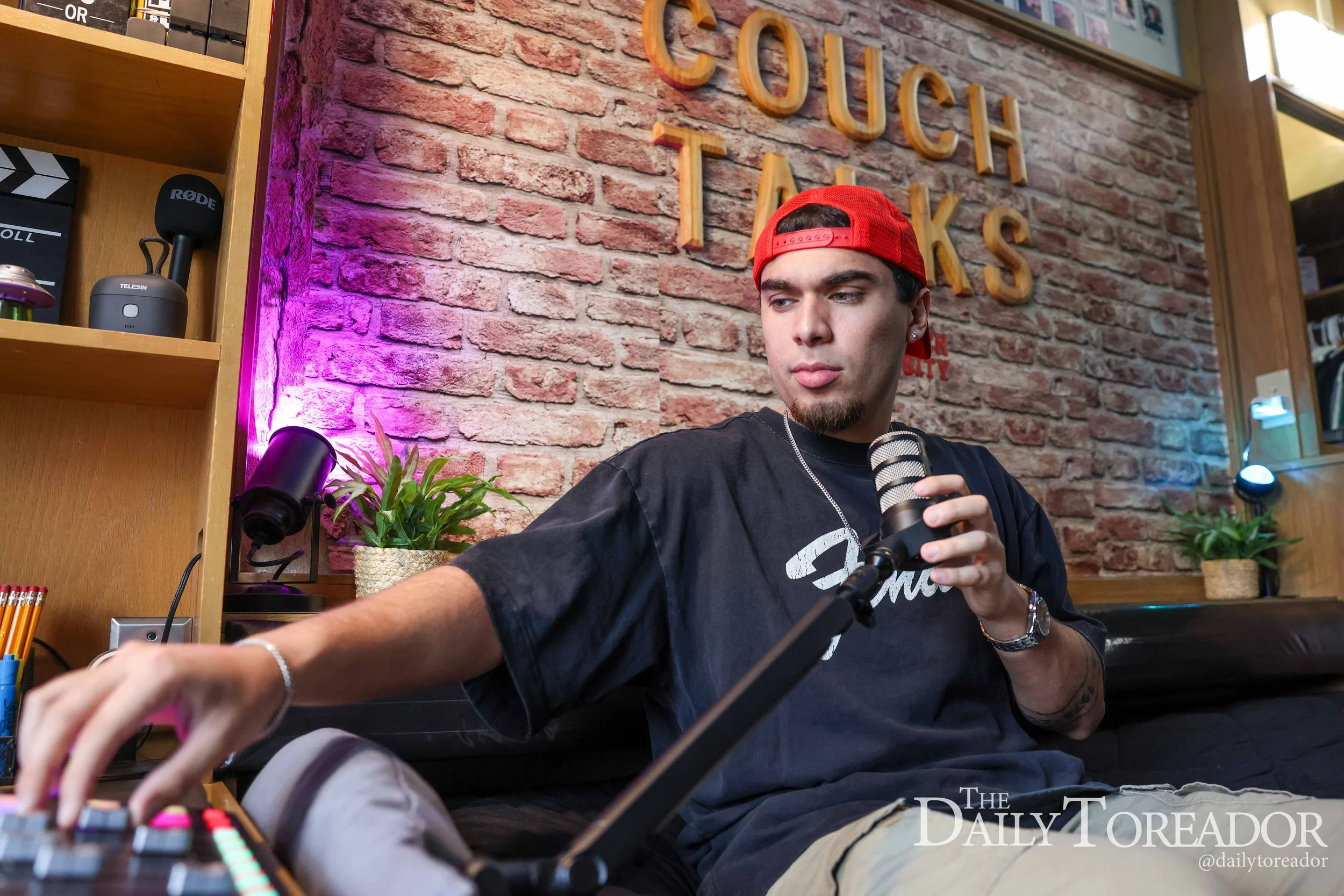 A young man wearing a red baseball cap backwards and a black t-shirt is sitting at a table holding a microphone. The background features a brick wall with the words "COUCH TALKS" in large wooden letters, some framed photos, and shelves with electroni