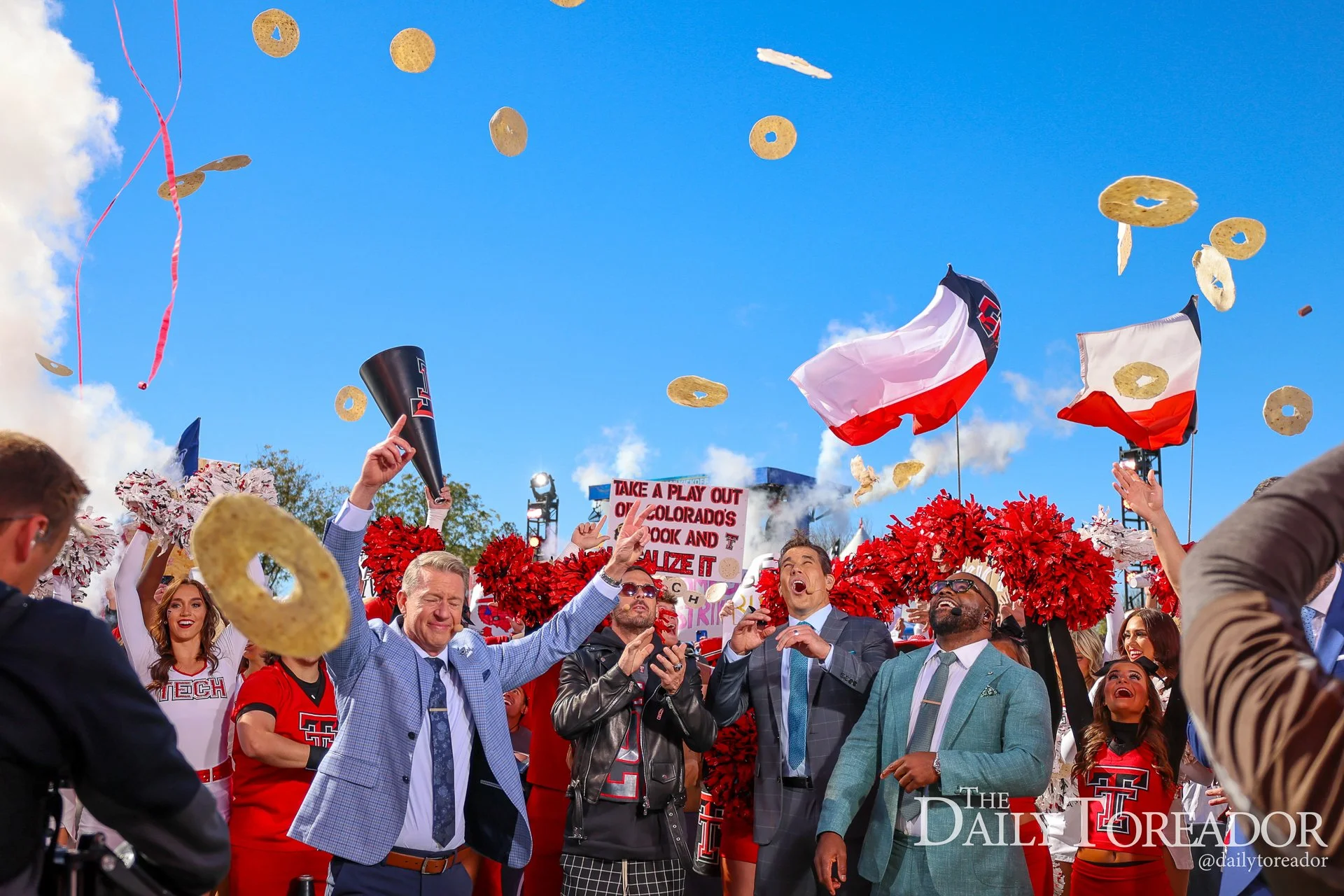 People celebrating at a college football game, waving flags and throwing cheerios into the air, with a clear blue sky above.