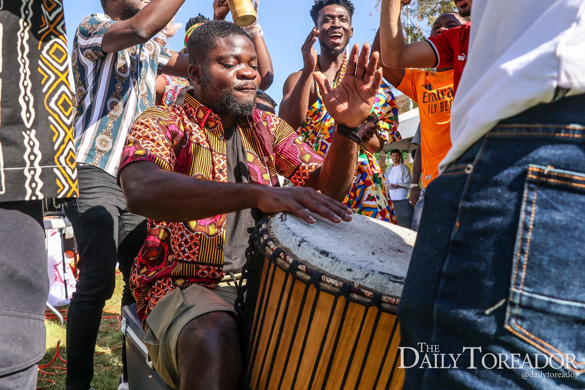 A group of people, mostly Black men, are gathered outdoors in a circle. One man in the center is sitting and playing a traditional drum, wearing a colorful patterned shirt. Others are smiling, clapping, and dancing, with some raising their hands, sug