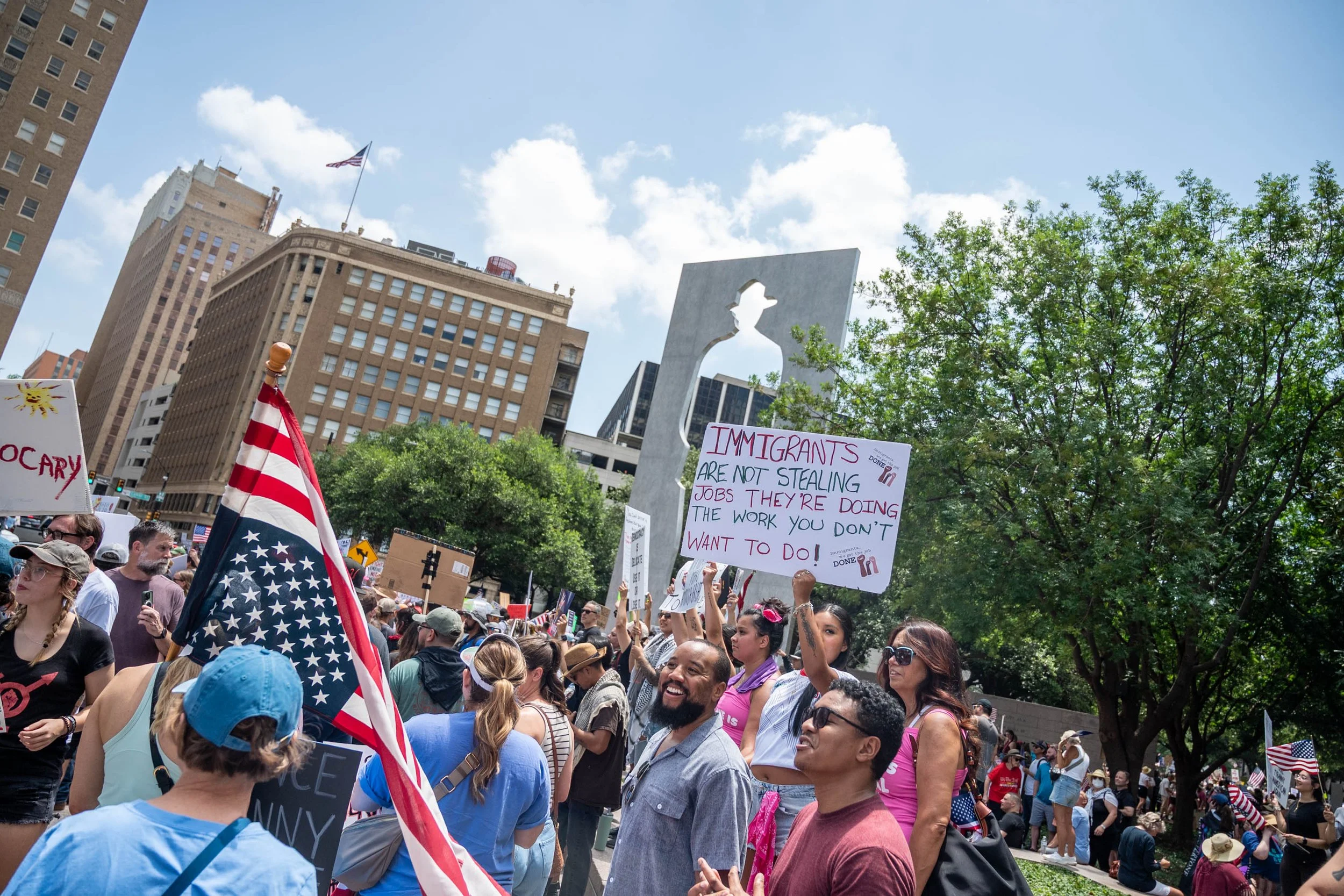 Crowd of protesters gathered in an outdoor urban area, holding various signs and American flags, with high-rise buildings and a blue sky with clouds in the background.