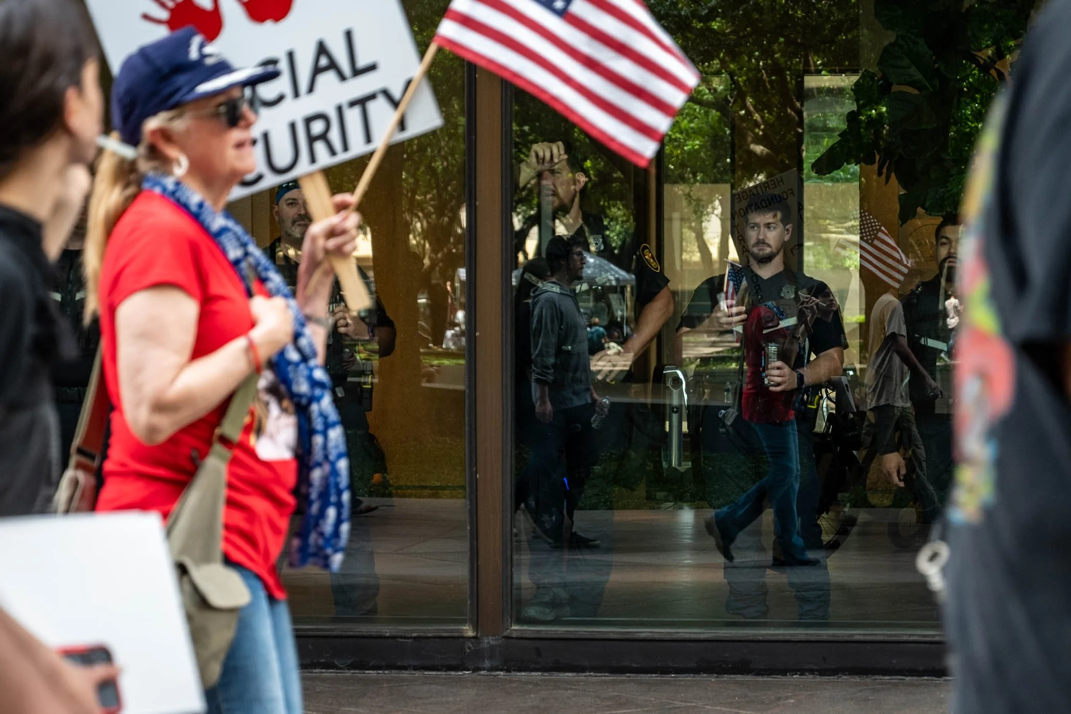 A woman in a red shirt holding a flag and a sign stands outside a building during a protest or demonstration, with police officers visible through a glass door reflecting the scene.