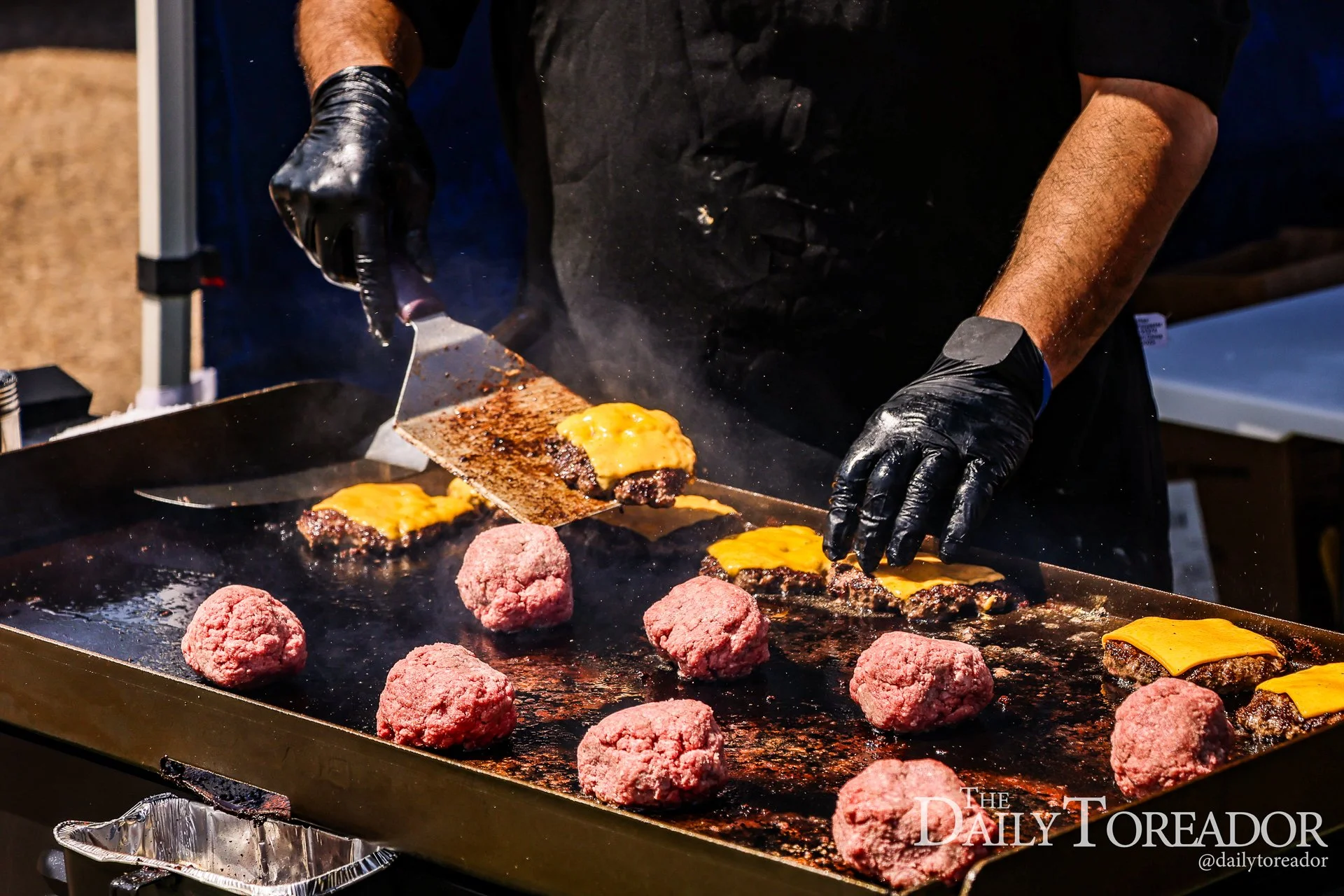 A person wearing black gloves cooking cheeseburgers and meatballs on a flat-top griddle.